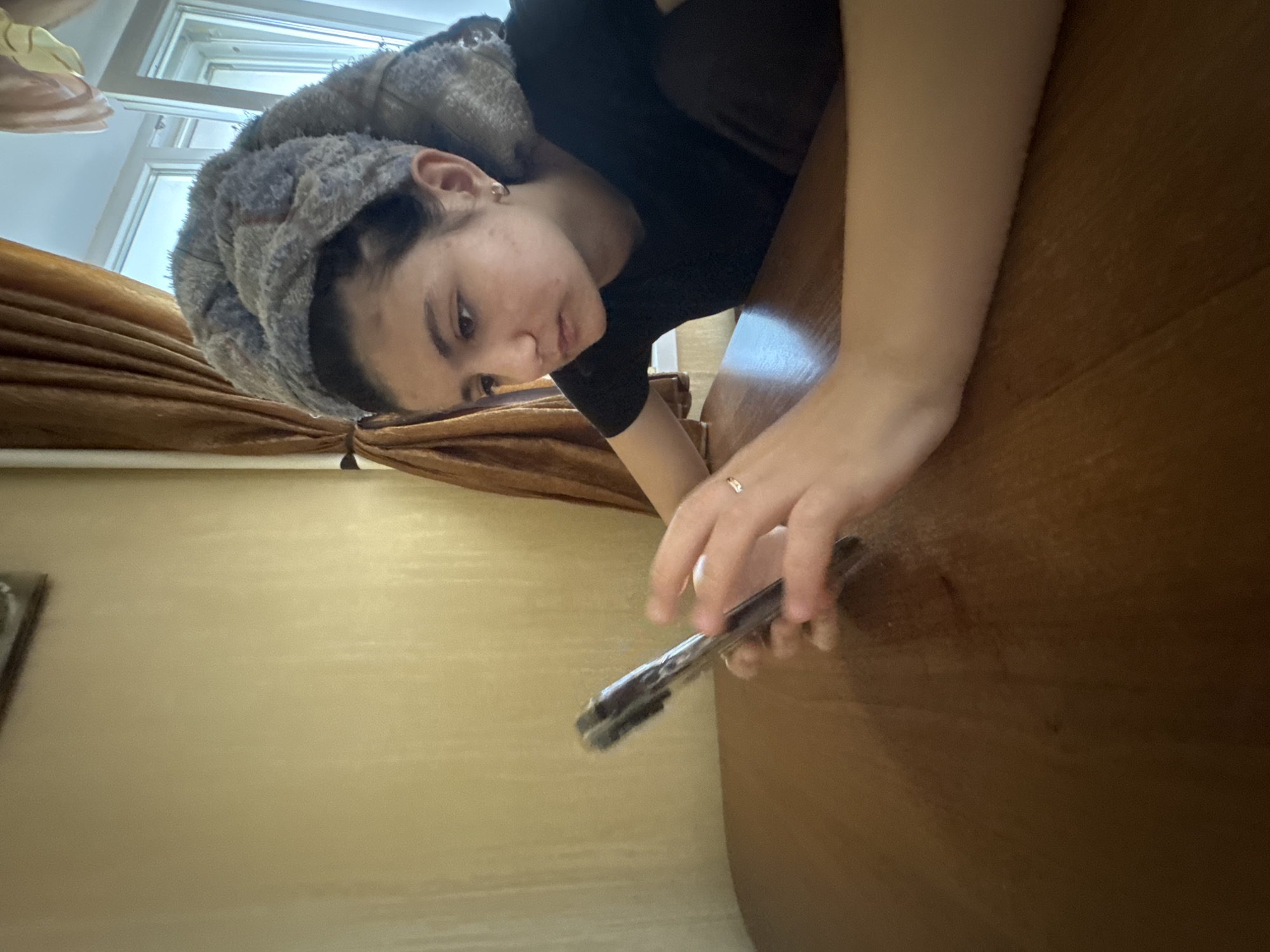 A young person is depicted in a close-up, rotated image, appearing to lie down or lean over a smooth, brown wooden surface. Their head is wrapped in a grey, fluffy towel, suggesting they may have recently washed their hair. They are wearing a simple black short-sleeved top. The person's left hand, adorned with a delicate gold ring on the ring finger, holds a smartphone, which they are looking at with a focused or neutral expression.
In the background, a richly textured, golden-brown curtain is visible on the left side, partially obscuring a bright window with white frames. A light beige or cream-colored wall can be seen further in the background. The natural light filtering through indicates it is likely daytime indoors. The setting suggests a casual, domestic environment, possibly a home. No visible text is present in the image. The photograph is taken from an unusual, rotated angle, giving a unique perspective on the scene.