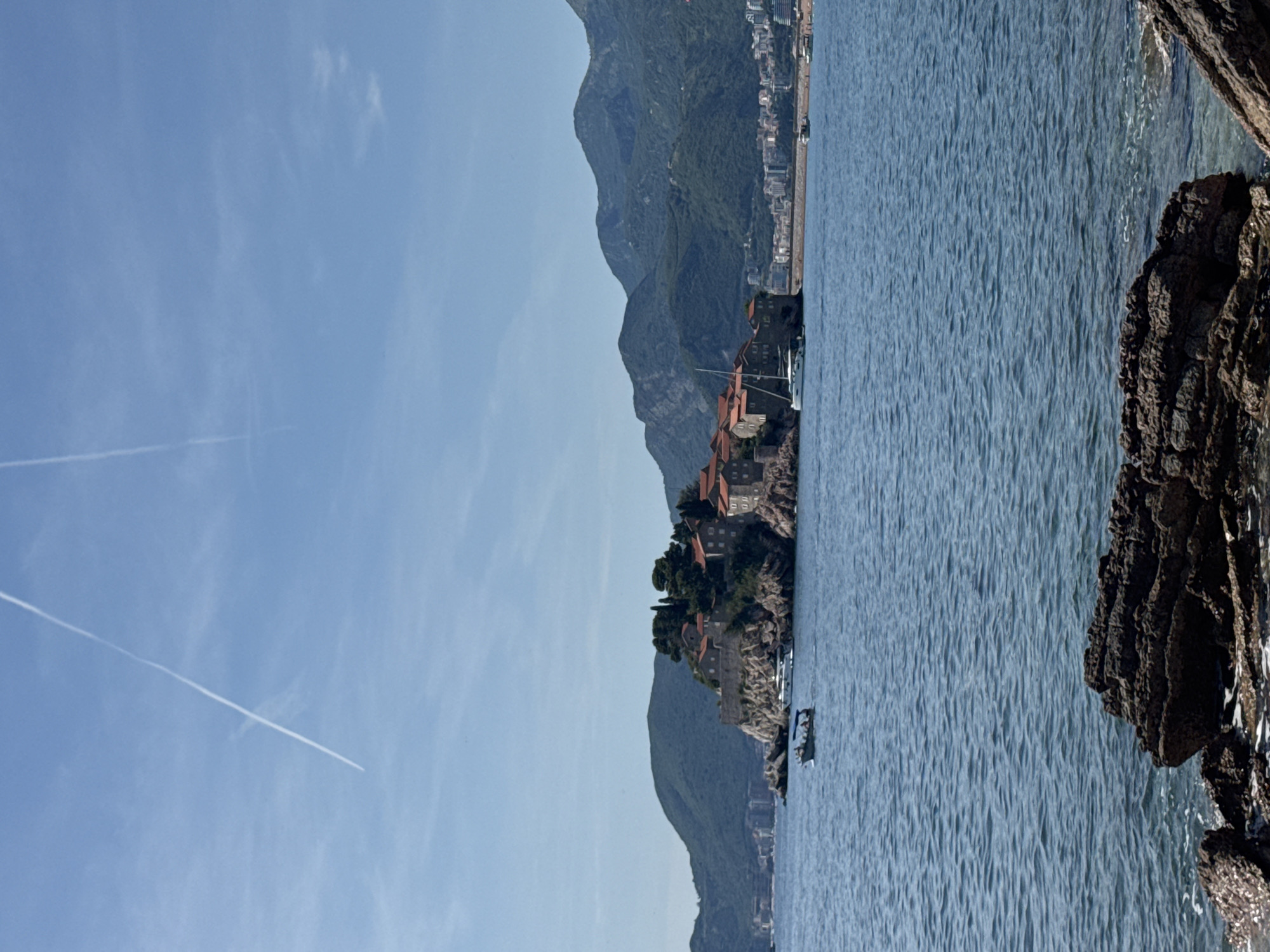The image depicts Sveti Stefan, Montenegro, on a clear day with a light blue sky. Two distinct white contrails intersect in the upper left quadrant of the sky. In the mid-ground, the fortified island of Sveti Stefan is prominent, covered with numerous terracotta-roofed stone buildings and scattered green trees. The island's rocky base extends into the water. To the left of the island, a small motorboat carries approximately five individuals. A large white yacht is moored on the left side of the island, near the buildings. On the right side of the island, another white sailboat or catamaran is docked.
Behind the island, a range of dark green, rugged mountains stretches across the horizon. To the right of the island, a narrow causeway connects it to the mainland, where a populated coastal area with multiple buildings and a beach is visible in the distant mid-ground. The sea in the foreground and mid-ground is a deep blue, exhibiting small ripples indicative of calm water. In the immediate foreground, dark, textured rock formations are partially submerged in the water, with clear water visible around them.