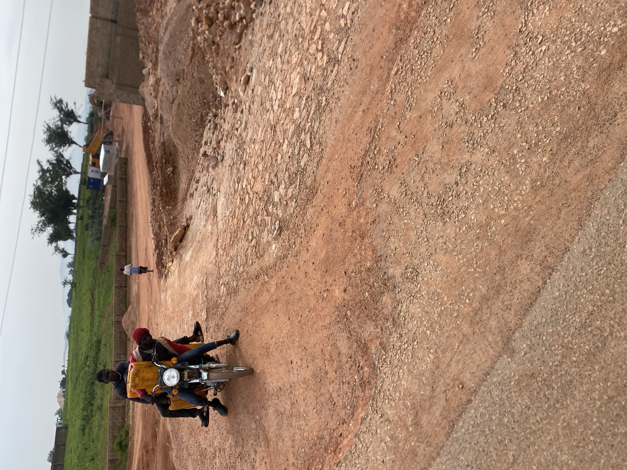 Two young men are riding on a motorcycle in Reifild-Ist, Nigeria. The man in front is wearing a dark long-sleeved shirt and blue jeans, while the man behind him is wearing a red cap and a red and black patterned shirt with blue jeans. They are carrying two large yellow plastic containers on the motorcycle. The motorcycle is red and has a headlight. The men appear to be on a dirt road with construction in the background. The environment is dusty and arid with some green vegetation in the distance. The construction site includes piles of dirt, bricks, and heavy machinery. Another person is visible riding a bicycle in the background.