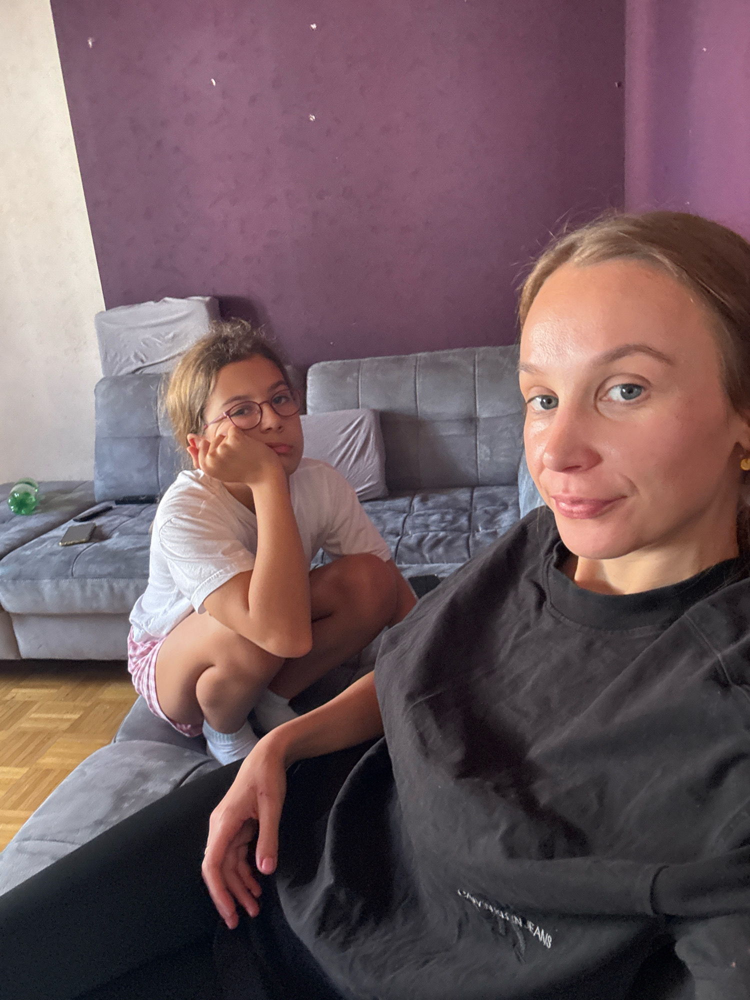The image depicts an indoor scene, likely a living room, with two individuals. A young girl with glasses is crouched on a gray sofa, resting her chin on her hand and looking bored. She is wearing a white t-shirt and pink and white striped shorts. To her right, a woman is taking a selfie, smiling at the camera. She is wearing a black t-shirt. The background features a purple textured wall and more sections of the gray sofa. Furniture and personal items like a phone and a green bottle are visible on the sofa. The lighting suggests an indoor setting during the daytime. The city of Überherrn, Germany, is not discernible from the visual elements in the image.