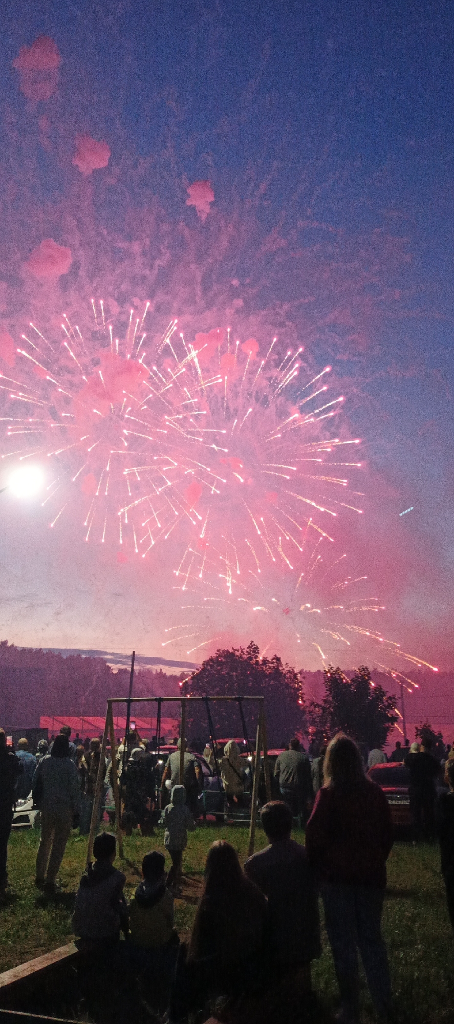 The image depicts a crowd of people gathered outdoors, watching a fireworks display in the twilight sky. The fireworks are predominantly pink and white, creating a vibrant spectacle against the deep blue and purple hues of the evening sky. In the foreground, several individuals are visible, some seated and others standing, facing away from the camera towards the fireworks. Behind them, more people are scattered across a grassy area, with some cars parked in the background. A wooden swing set structure is partially visible in the middle ground. The overall atmosphere suggests a festive event or celebration taking place in an open, outdoor venue, possibly a park or a field, on the outskirts of the city of Chaikovsky, Russia.