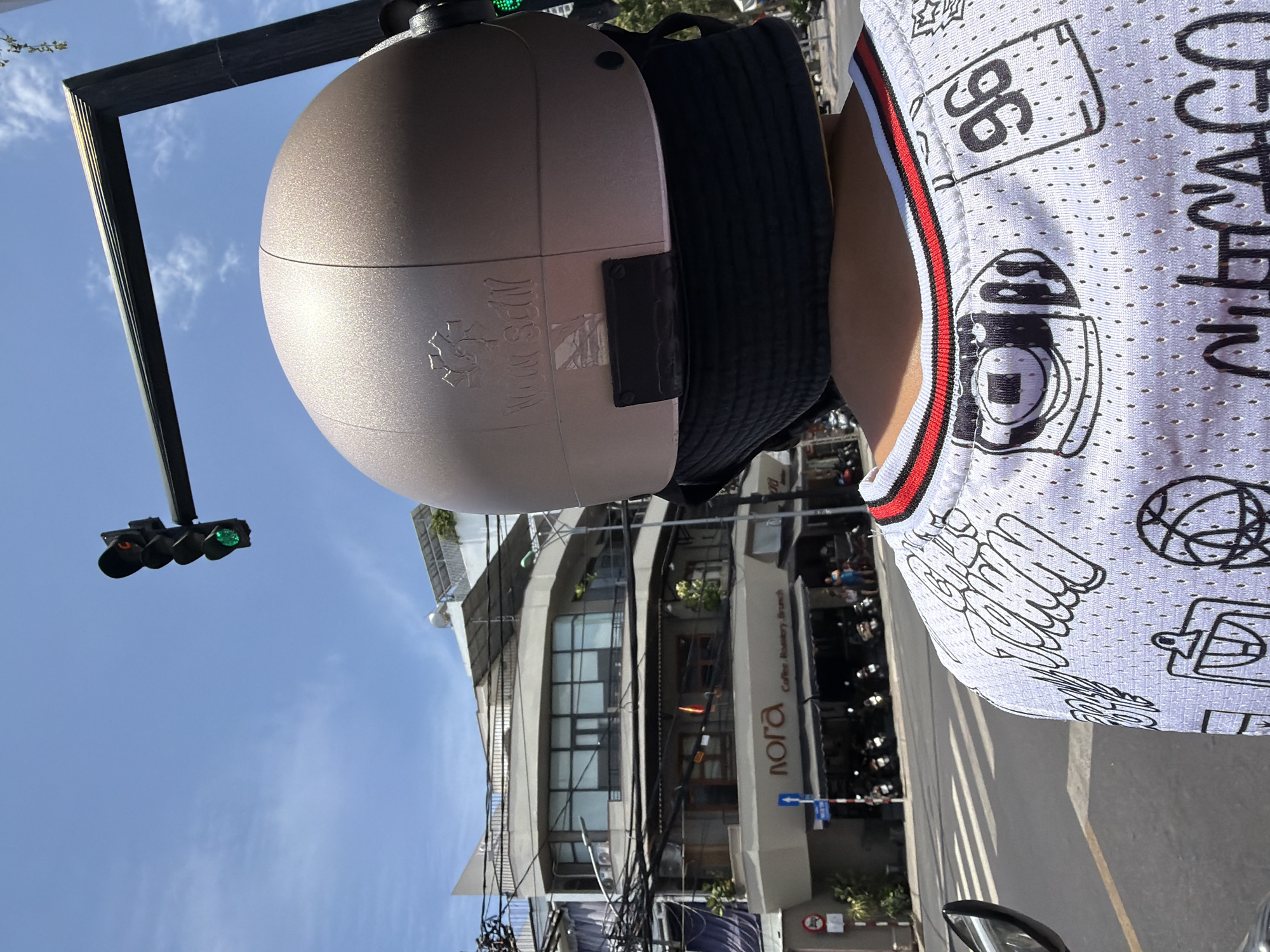 In Nha Trang, Vietnam, this image captures a street scene from a first-person perspective, likely from the back of a motorcycle. The foreground is dominated by the back of a person wearing a light-colored helmet and a white mesh jersey with black graphic prints, including the number "96" and the word "CHICAGO" in stylized lettering. The jersey has a red and black striped trim around the neckline. A black fabric is visible under the helmet, suggesting it's part of a mask or head covering.
Dominating the background is a building with a curved facade, featuring multiple levels with large windows and balconies. A sign on the lower level reads "nora Coffee. Roastery. Branch." A traffic light is prominently displayed in the upper right portion of the image, showing a green light. Wires and cables crisscross above the street, a common sight in Vietnamese cities. The street below shows a pedestrian crossing and parked motorcycles, indicating a busy urban environment. The sky is blue with scattered white clouds.
