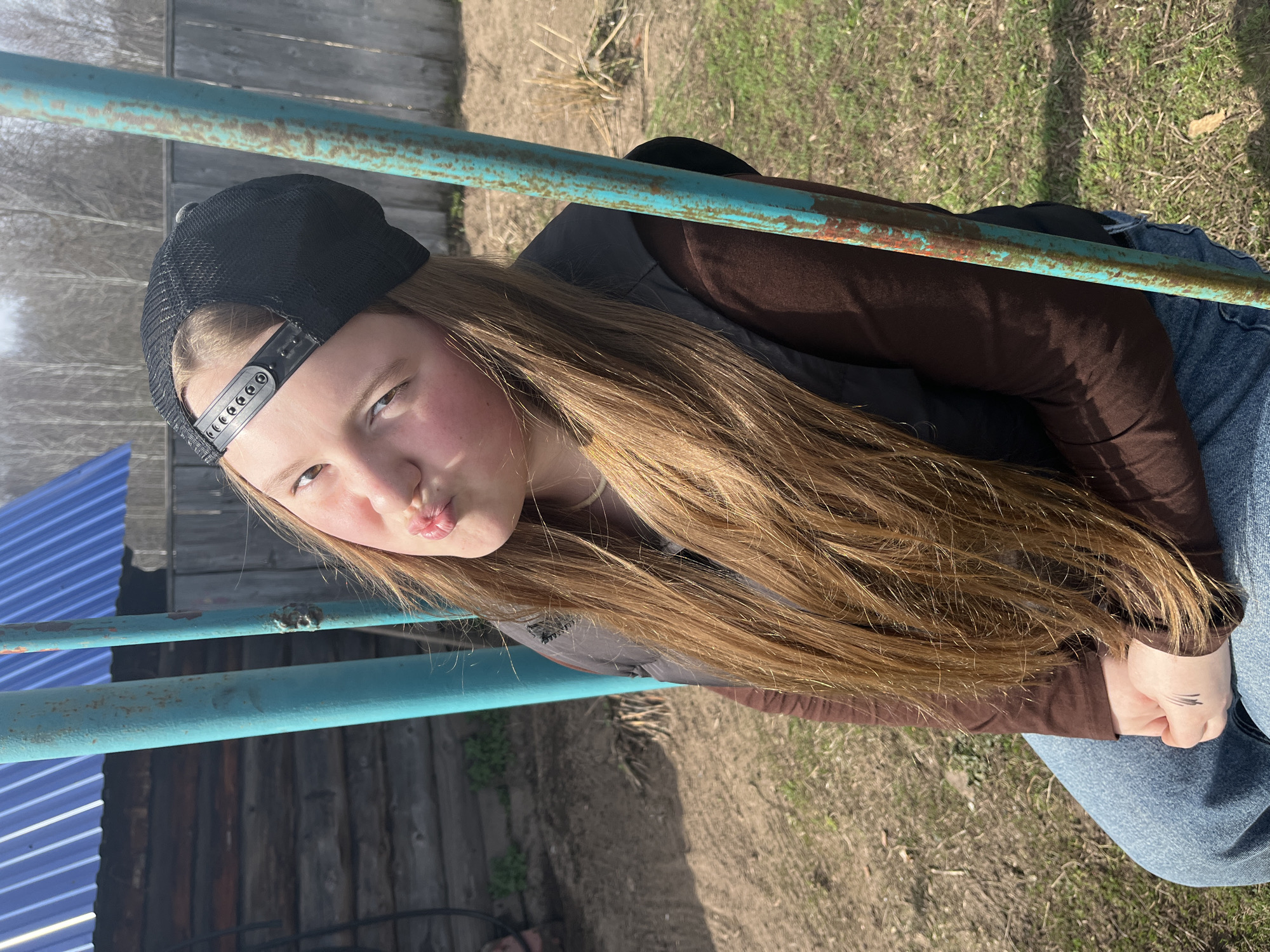 A young person, likely female, poses outdoors in daylight. The individual wears a black baseball cap, a brown long-sleeved shirt layered under a black vest, and blue jeans. They are seated, positioned behind teal-painted metal bars with rust. The background includes a wooden fence, a structure with a blue corrugated roof, and patches of dry grass and sparse vegetation. The individual is making a kissing face towards the camera. There are marks on the person's hand. The setting appears to be in a rural or semi-rural location. Considering the location is in Буренка, Russia, the environment likely reflects a typical Russian village or countryside.