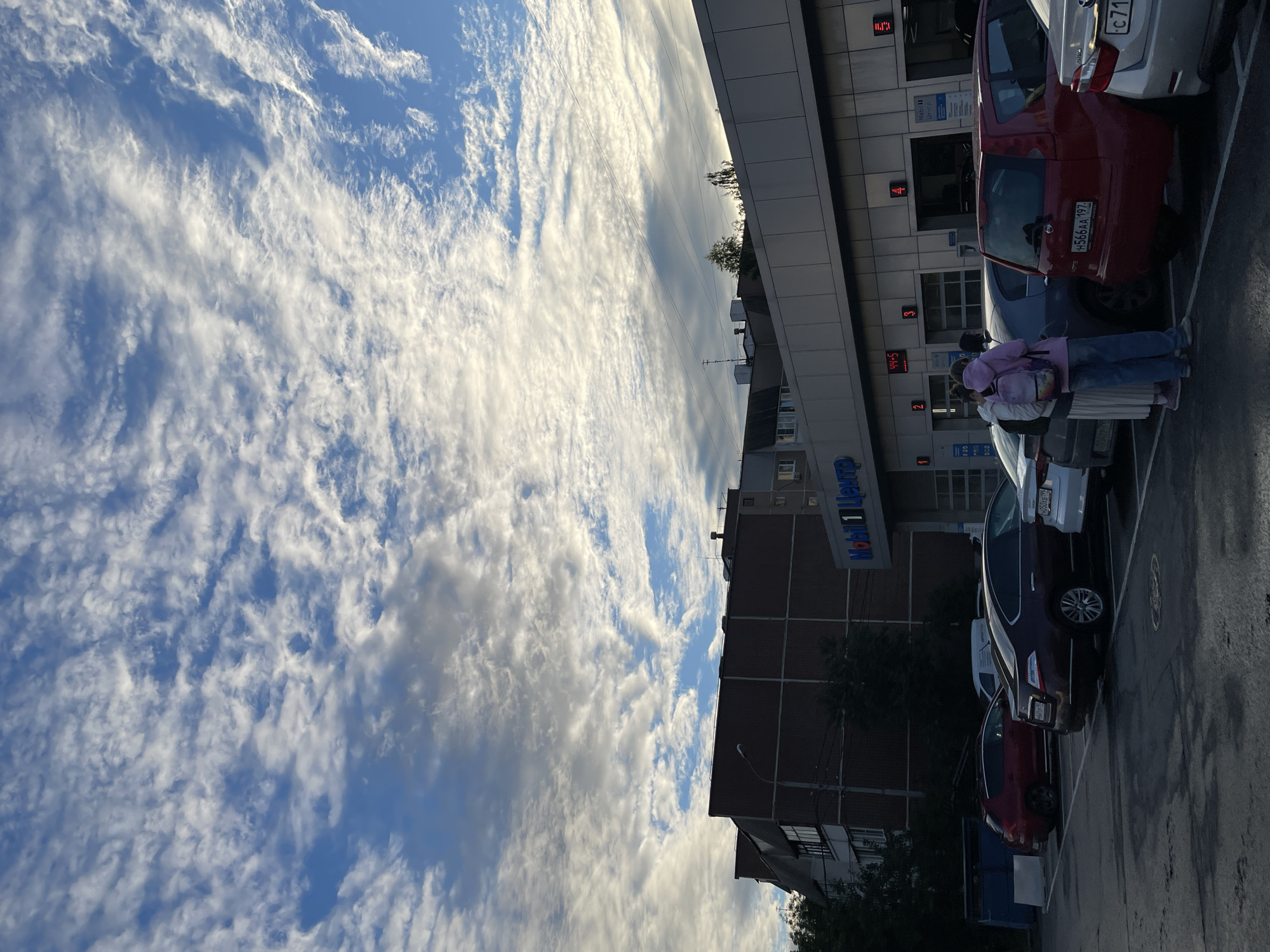 This image captures a parking lot scene with a modern building and a vast sky filled with dynamic clouds. The sky is a prominent feature, with wispy, white clouds streaking across a vibrant blue expanse, suggesting a bright and possibly windy day. The sun is low in the sky, casting long shadows and illuminating the clouds from the side, indicating it's either early morning or late afternoon.
In the foreground, several cars are parked. A red hatchback is visible, with a license plate that reads "h566AA 197". A dark-colored car is parked next to it, followed by a white car. A person wearing a pink hoodie and jeans, with a backpack, is standing near the red car.
The building in the background appears to be a commercial structure with a modern facade, featuring glass windows and metallic paneling. One sign on the building reads "Mobil 1 Центр" in blue letters. Another sign displays digital numbers, possibly indicating time or service information, with digits like "11.11" and "44.5" visible.
The overall scene is likely a public parking area adjacent to a business establishment in Moscow, Russia, during daylight hours with partly cloudy weather. The lighting suggests a time of day with a warm, diffused light, and the presence of cars and a building points to an urban or suburban setting.