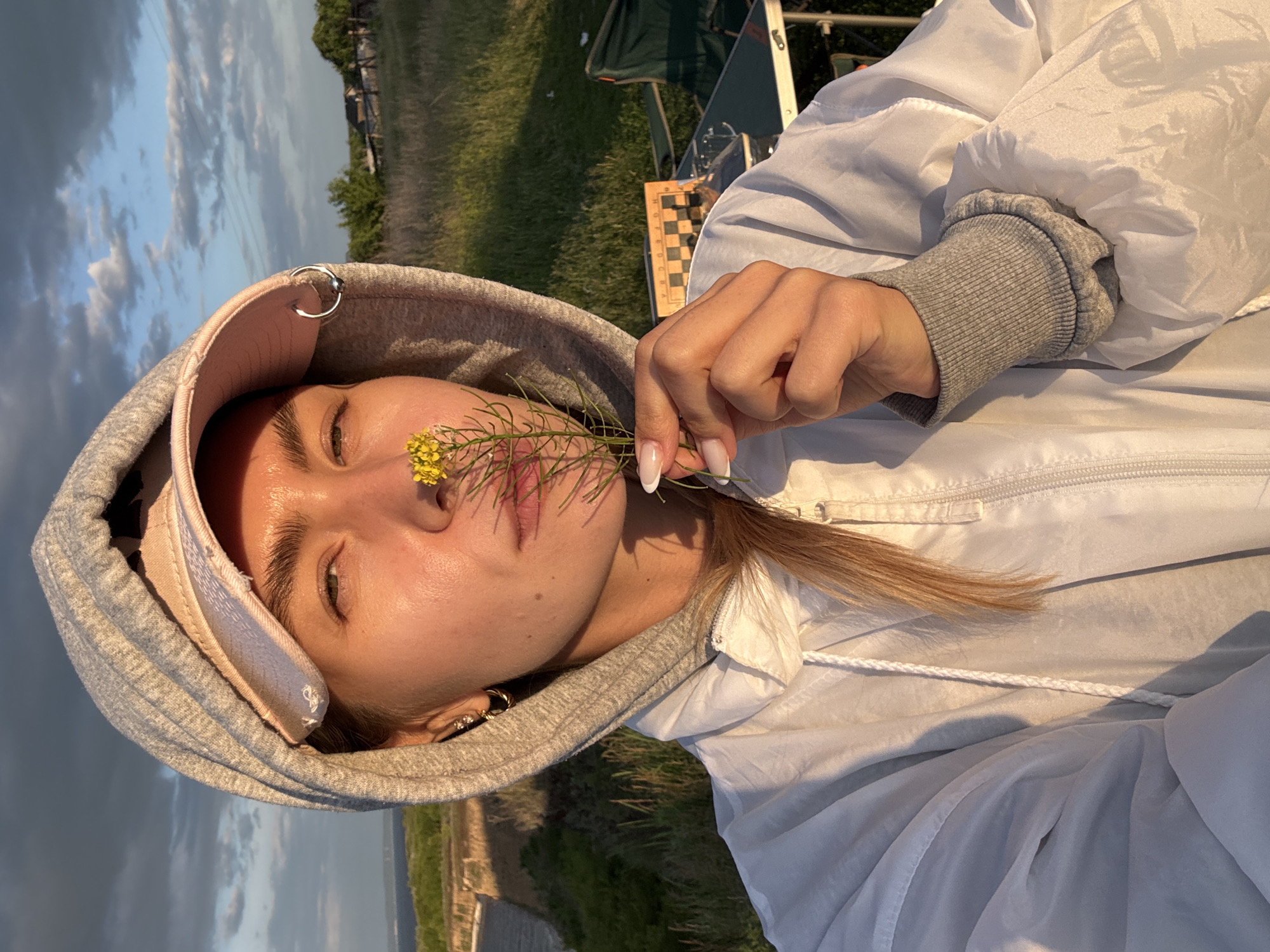 A female subject is captured in a close-up, waist-up selfie, illuminated by the warm, golden light of either sunrise or sunset. She wears a light grey hooded sweatshirt with the hood up, a pink baseball cap beneath the hood, and a white windbreaker jacket over the sweatshirt. A silver hoop earring and a small stud are visible in her left ear, while a silver ring piercing is attached to the edge of her hood. Her left hand, featuring nails with white tips (French manicure), holds a sprig of yellow wildflowers to her face, positioned between her nose and mouth. Her eyes are partially squinted, directed towards the camera.
The setting is an outdoor, natural environment. To the left, a large body of water, likely a river or lake, extends towards a distant landmass on the horizon. The sky above presents a mix of dark grey clouds and lighter patches with white cumulus clouds. The immediate foreground consists of dry, sun-drenched grass. To the right, a grassy field extends to distant trees and structures indicative of a rural settlement. A green folding table is visible in the mid-ground, displaying a wooden, folded chessboard and partially obscured green camping chairs, suggesting a leisure or camping activity. The light casts distinct, long shadows.