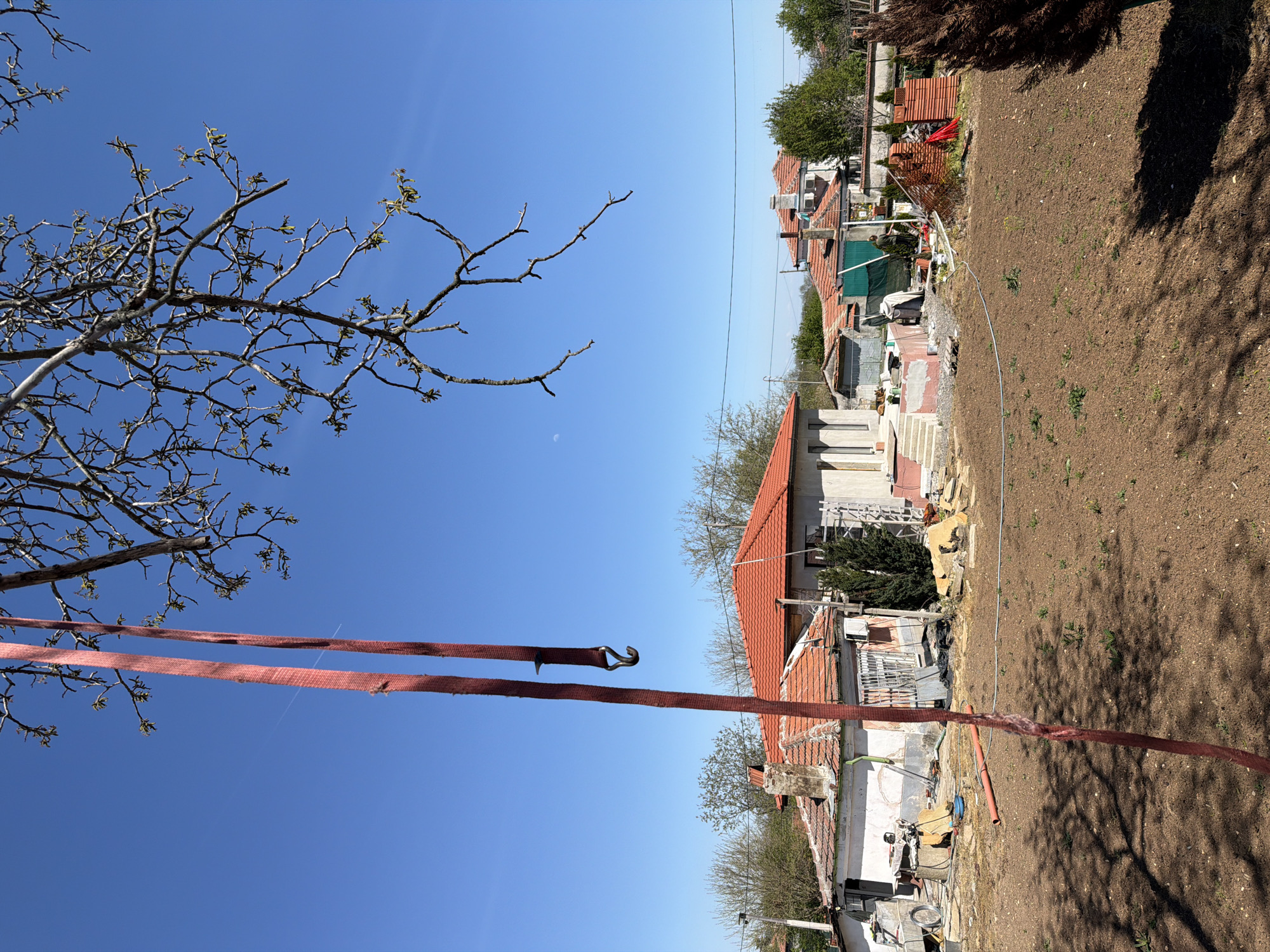 The image shows a residential area on a clear day, with a focus on a building undergoing construction or renovation. The foreground includes a bare tree with new buds, and a red nylon strap or rope is stretched across the scene. In the background, several houses with red tile roofs are visible, interspersed with trees and other vegetation. The ground is mostly bare soil, suggesting a garden or construction area. There are no people visible in the image.
