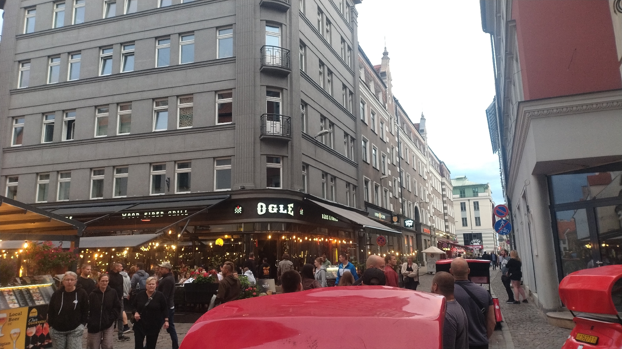 In Riga, Latvia, a bustling street scene unfolds with a multi-story building dominating the left side of the frame. The building, painted a muted grey, features numerous windows and a few balconies. Below the building, a covered outdoor seating area for a restaurant is illuminated by strings of lights, creating a warm and inviting atmosphere. Several people are visible, some standing and talking, others walking, and some are seated at tables under the awning. The restaurant's signage, featuring the name "OGLE", is prominent. A red vehicle, possibly a scooter or a small car, is parked in the foreground, partially obscuring the view of the street. The right side of the image shows another building with a pink facade, and further down the street, other buildings and pedestrians are visible. The sky is overcast, suggesting a cloudy day.