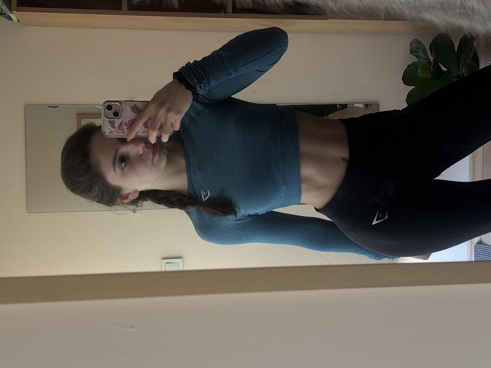 A woman is taking a mirror selfie indoors. She is wearing a teal long-sleeved crop top and black leggings with the Gymshark logo on the left thigh. Her dark hair is braided. She is holding a smartphone with a patterned case. A mirror reflects her image and part of the room, which includes a shelf, a plant, and a light switch. The location is likely indoors, but there is not enough information to determine if it is in the city of Varna, Bulgaria.