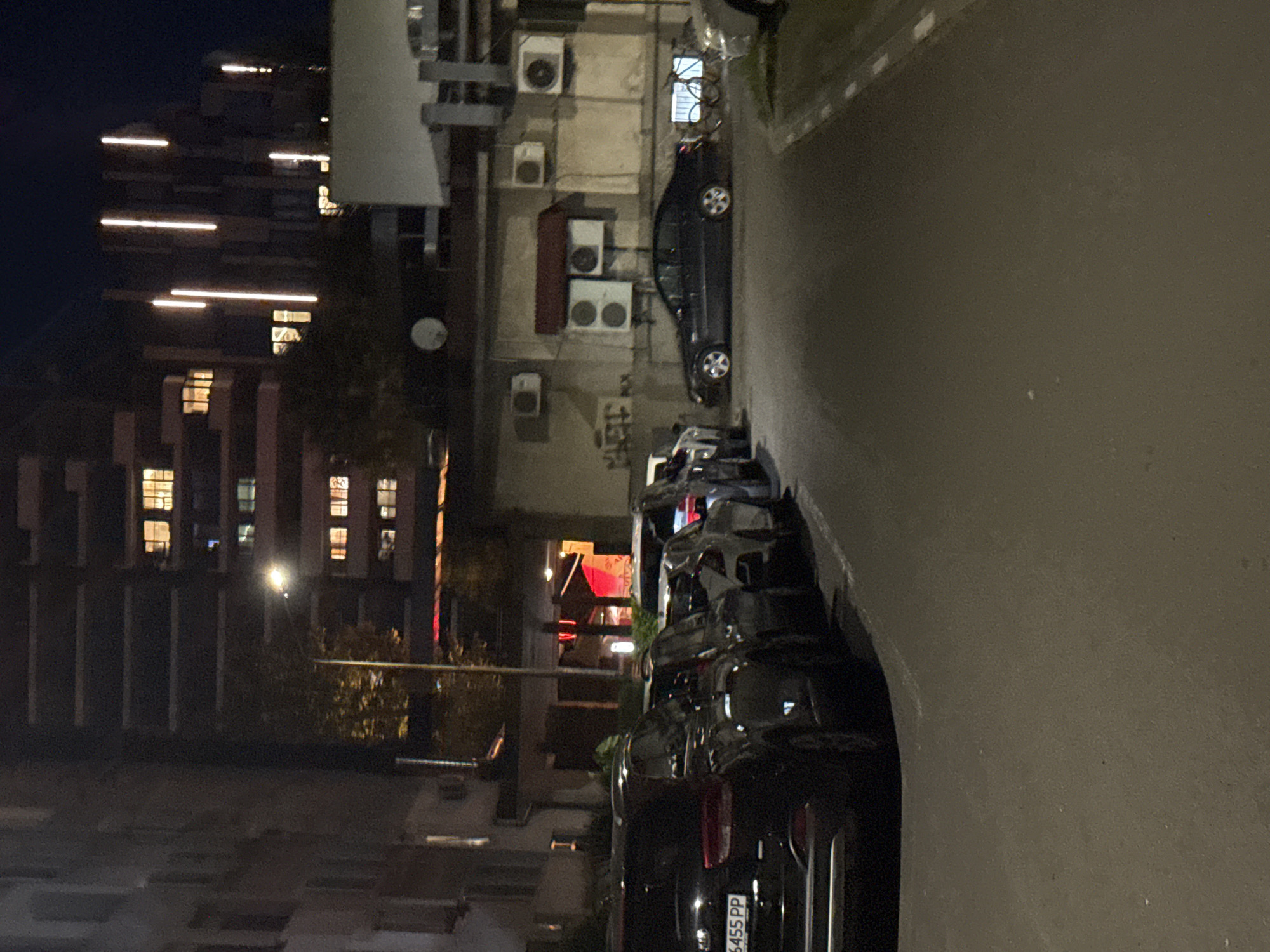 This is a nighttime street-level photograph taken in the city of Burgas, Bulgaria. The scene depicts a row of parked cars on a paved street. In the background, there are multi-story buildings with illuminated windows, suggesting a residential or commercial area. One of the buildings features a distinctive modern architectural style with vertical light fixtures. Several air conditioning units are visible mounted on the exterior walls of a building on the right side of the image. A street lamp with a bright light is also present, illuminating part of the street and trees. The overall impression is of a quiet urban environment at night.