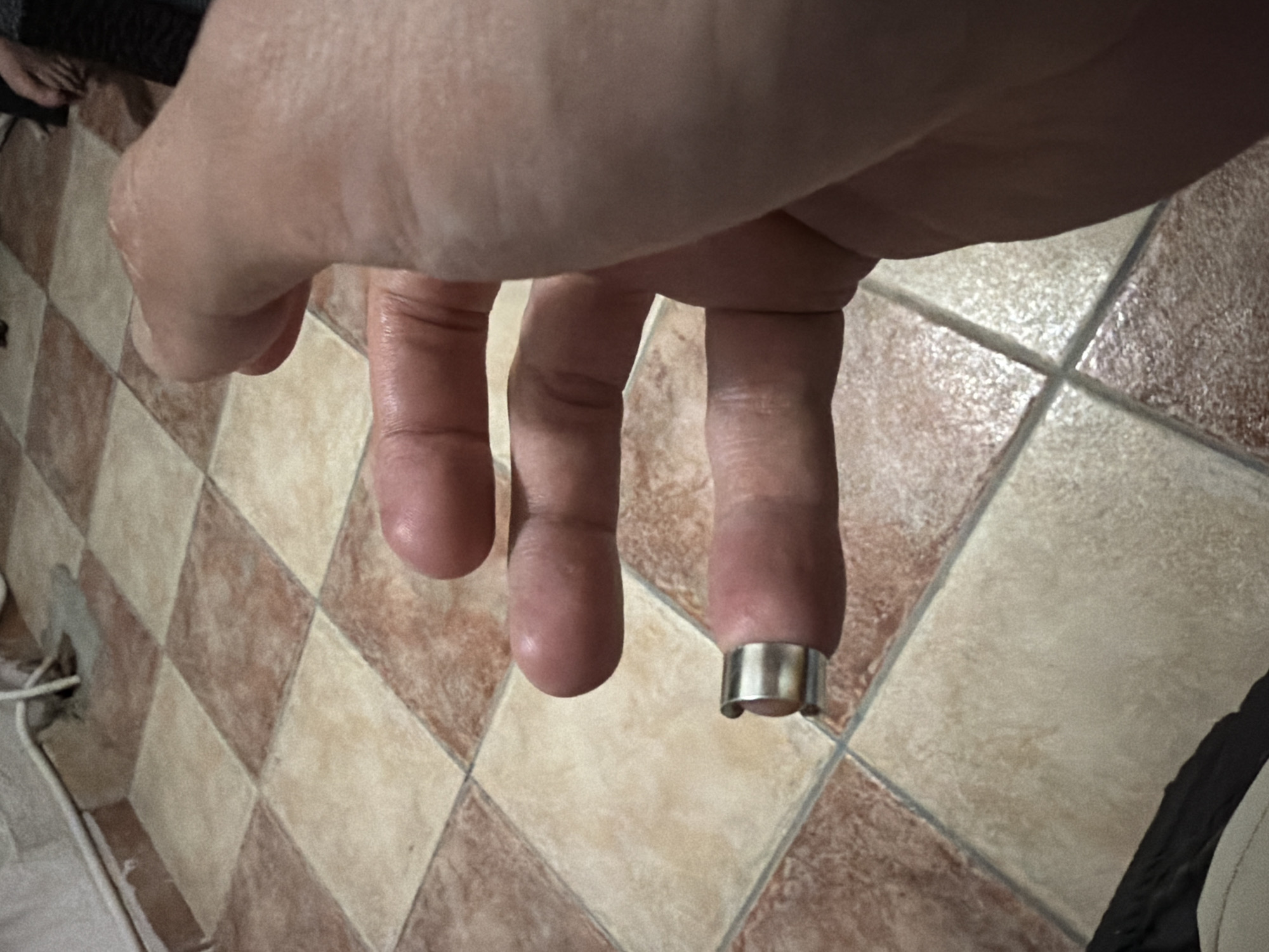 A close-up shot shows a hand extending towards a patterned tile floor. The index finger has a wide silver ring on it. The background floor consists of alternating beige and brown square tiles. A foot is slightly visible at the upper left corner, near a black object. An electrical cord protrudes from the floor along the wall at the lower-left. A portion of a dark object with beige trim is partially visible at the lower right. The image does not contain sufficient information to determine if this location is in Budva, Montenegro.
