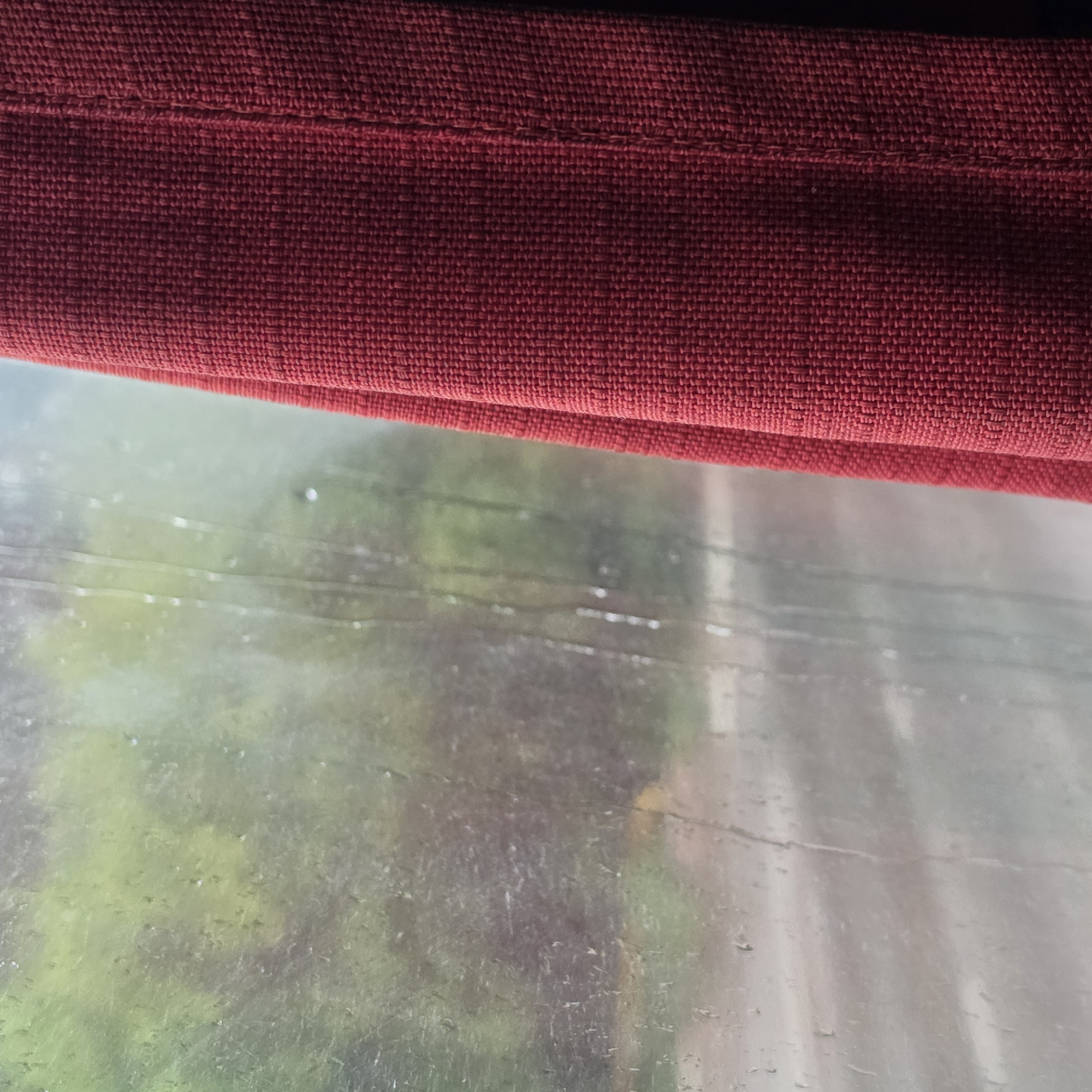 The image presents a view through a wet window, partially obscured on the right by a red curtain. This scene is located in the city of Chaikovsky, Russia. The window glass is covered with numerous water droplets and vertical streaks, indicating rain or heavy condensation. Beyond the window, the exterior environment is blurry and indistinct. The upper and central left portion reveals a muted expanse of green and dark grey, consistent with dense foliage or trees under overcast conditions. Below this, a wide, flat, grey-toned area suggests a wet road or paved surface, also blurred by the water on the glass and the ambient conditions. No specific features like vehicles, buildings, or street markings are discernible in the exterior. The lighting outside appears soft and diffused, indicative of an overcast sky. On the right side, a deep red fabric curtain occupies approximately one-quarter of the frame. The curtain displays a clear, consistent woven texture, possibly a basketweave, with individual threads forming a uniform pattern. It is draped, exhibiting a prominent vertical fold or seam line that creates a subtle shadow down its length. No people or animals are visible within the frame.