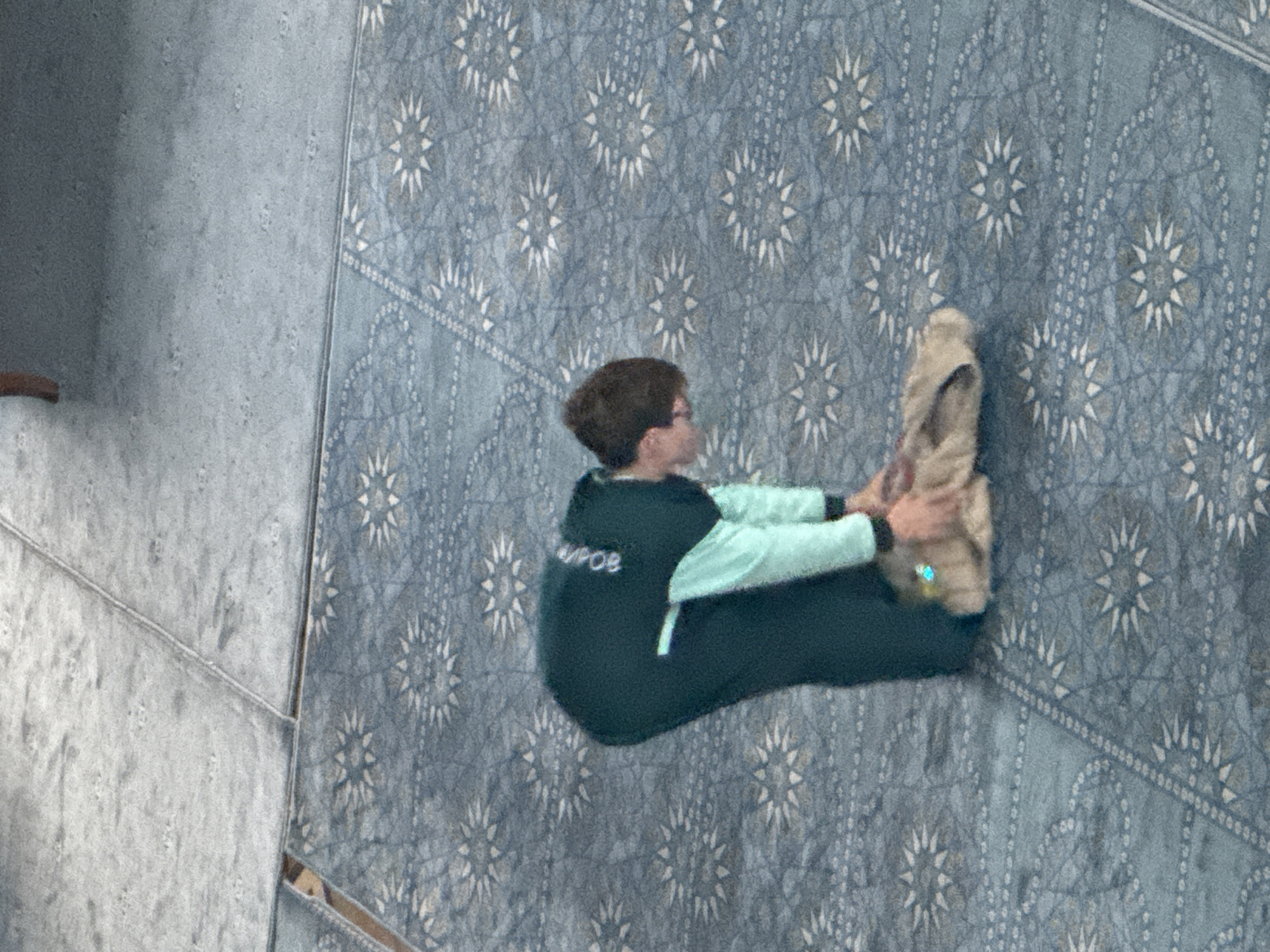 A person sits on the floor in Kazan, Russia. The person is wearing a dark green jumpsuit with light green sleeves and is leaning forward, holding a beige object. The floor is covered with a patterned carpet in shades of blue and gray. To the left is a gray wall or floor with a seam.