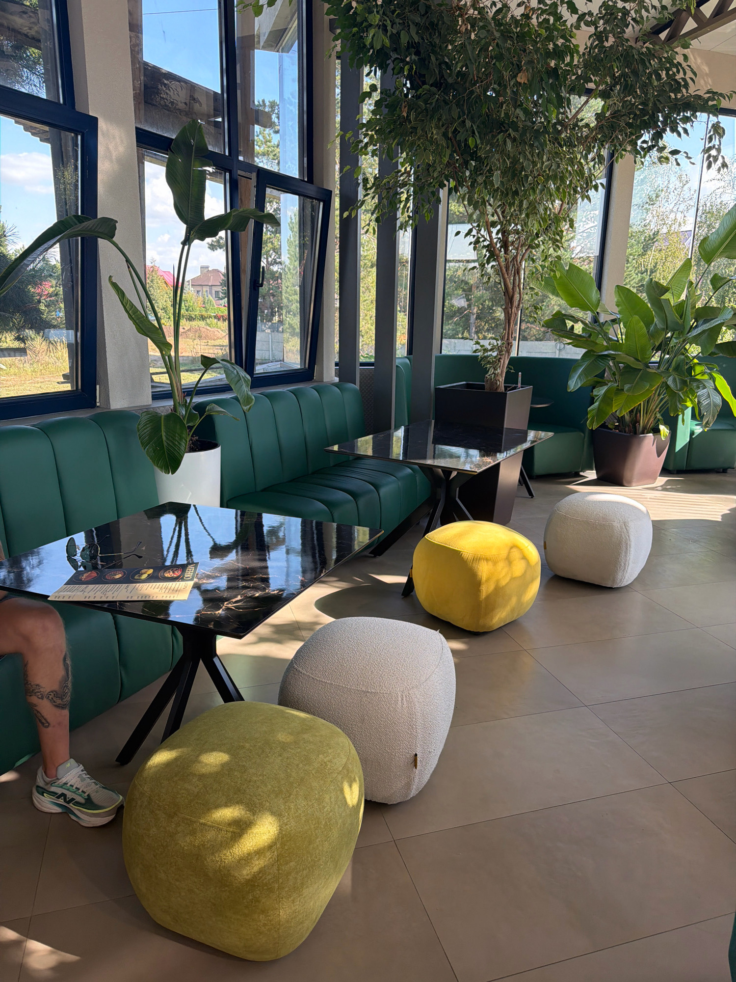 The image depicts the interior of a modern cafe in Malaya Tsarevschina, Russia. The cafe features a spacious seating area with large windows that offer a view of the outdoors. Inside, plush green booth seating lines the walls, accompanied by black marble-topped tables with black metal bases. On one table, a menu and eyeglasses are visible. Several plush ottomans in yellow and off-white are arranged on the tiled floor, serving as additional seating. Large indoor plants, including a ficus tree and a bird of paradise plant, are strategically placed throughout the space, adding a touch of nature to the contemporary design. The lighting suggests it is daytime, with natural light streaming through the windows. A person's lower leg and foot, wearing a sneaker, are visible on the left side of the frame, indicating their presence in the cafe. The overall impression is one of a clean, well-lit, and comfortable dining environment.