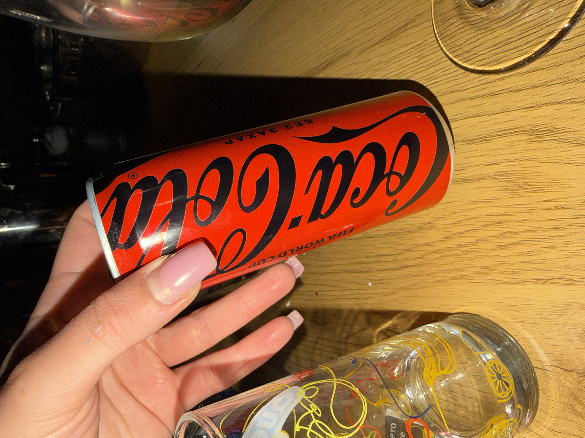 A hand with pink manicured nails holds a red Coca-Cola can. The can has the Coca-Cola logo in black script and text in Cyrillic script which reads "БЕЗ ЗАХАР" (sugar-free). The hand and can are placed over a wooden table. To the left of the can is a clear glass with yellow and red designs. The background is dark and out of focus, showing hints of other objects and a dark surface. The image appears to be taken indoors. There is no identifiable location in the city of Enina, Bulgaria.