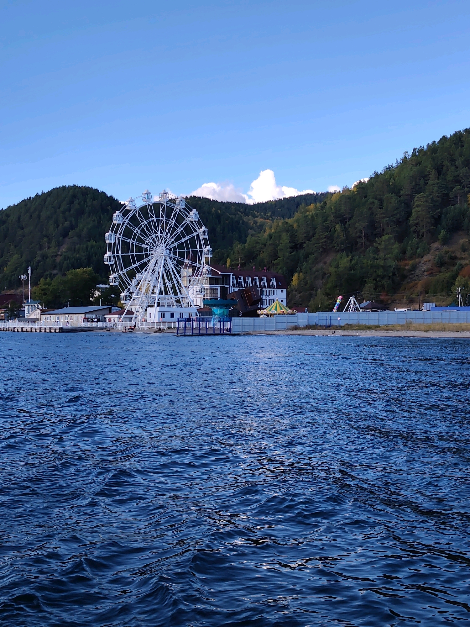 The image depicts a waterfront scene in Listvyanka, Russia, featuring a large white Ferris wheel as the prominent focal point. The Ferris wheel stands on a paved area adjacent to the water, with several buildings and other amusement park attractions visible in the background. The buildings appear to be part of a resort or entertainment complex, with a white facade and a red roof. To the right of the Ferris wheel, there is a smaller amusement ride with colorful elements. The background is dominated by a steep, forested hillside, indicative of the natural landscape surrounding Lake Baikal. The water in the foreground is a deep blue, showing small ripples and reflecting the light. The sky is clear and blue with a few wispy white clouds. The overall impression is of a tourist destination with recreational facilities situated by a large body of water and backed by natural scenery.