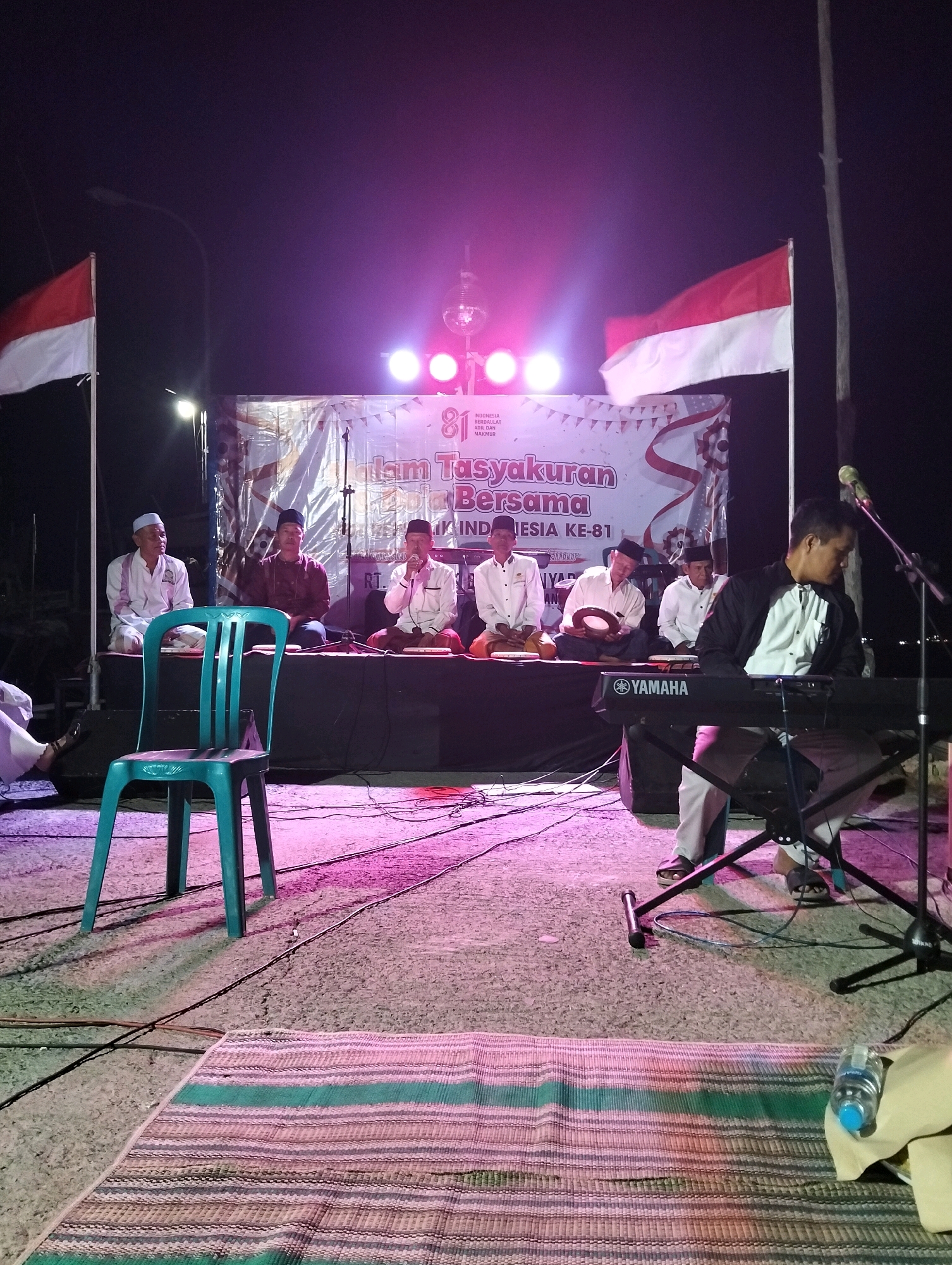 The image depicts an outdoor nighttime event in Paciran, Indonesia. A stage is set up with a banner that reads "Tasyakuran Bersama" and "INDONESIA KE-81". Several men are seated on the stage, some wearing traditional attire. One man is playing a keyboard, and another appears to be speaking into a microphone. Two Indonesian flags are visible on either side of the stage. In the foreground, a turquoise plastic chair is placed on a textured ground, with wires and lighting casting colorful shadows. A woven mat is laid out on the ground in front of the stage. The overall atmosphere suggests a cultural or celebratory gathering.