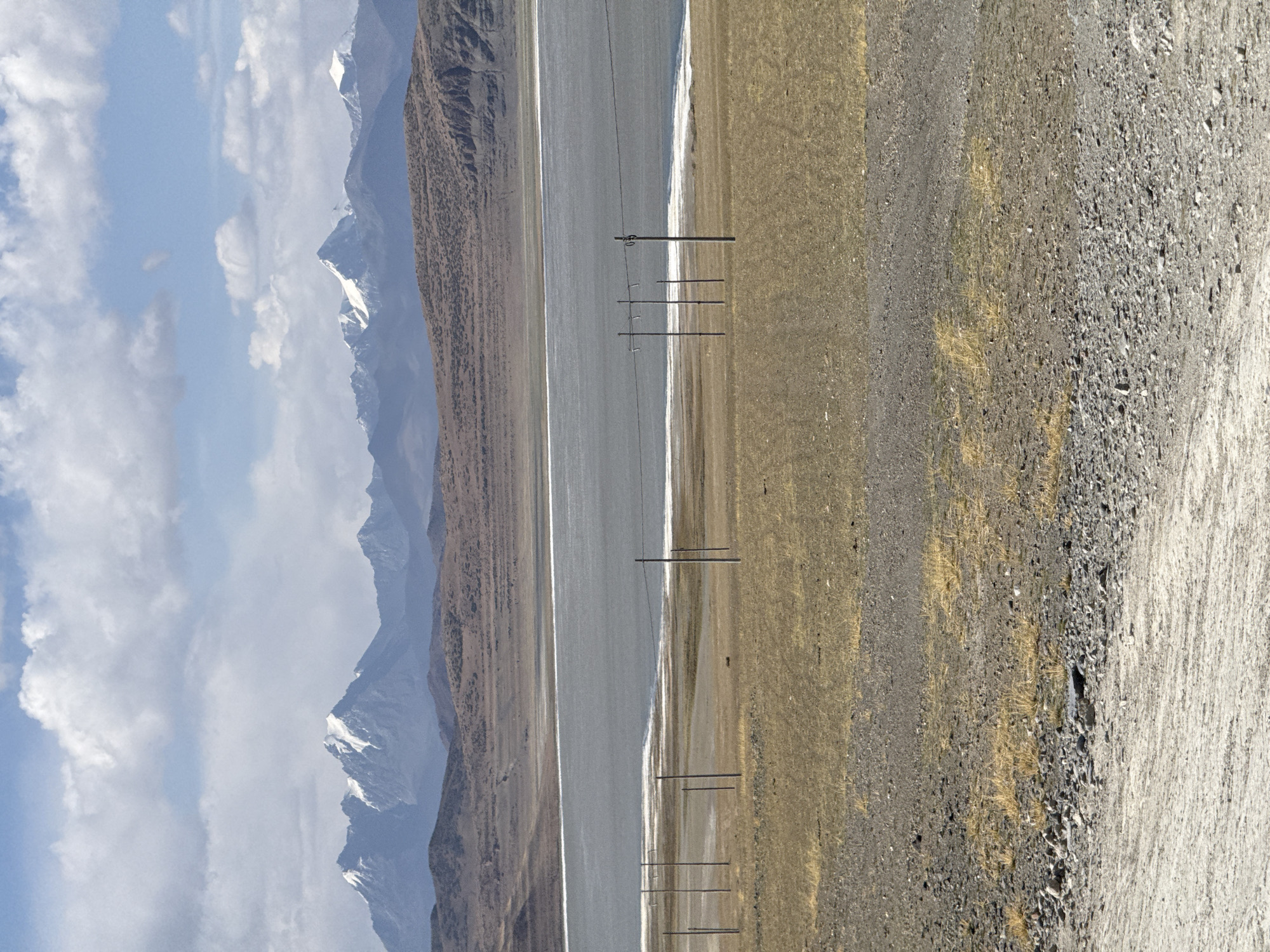 The image depicts a mountainscape alongside a paved road in Gyirong County, China. The foreground is composed of a gravel road, transitioning into a stretch of yellow grassland. Parallel to the grassland runs a paved road. Adjacent to the road, a mountainous landscape extends. The mountains in the background are capped with snow, while the sky is partially covered with clouds. Power line poles stand alongside the road.