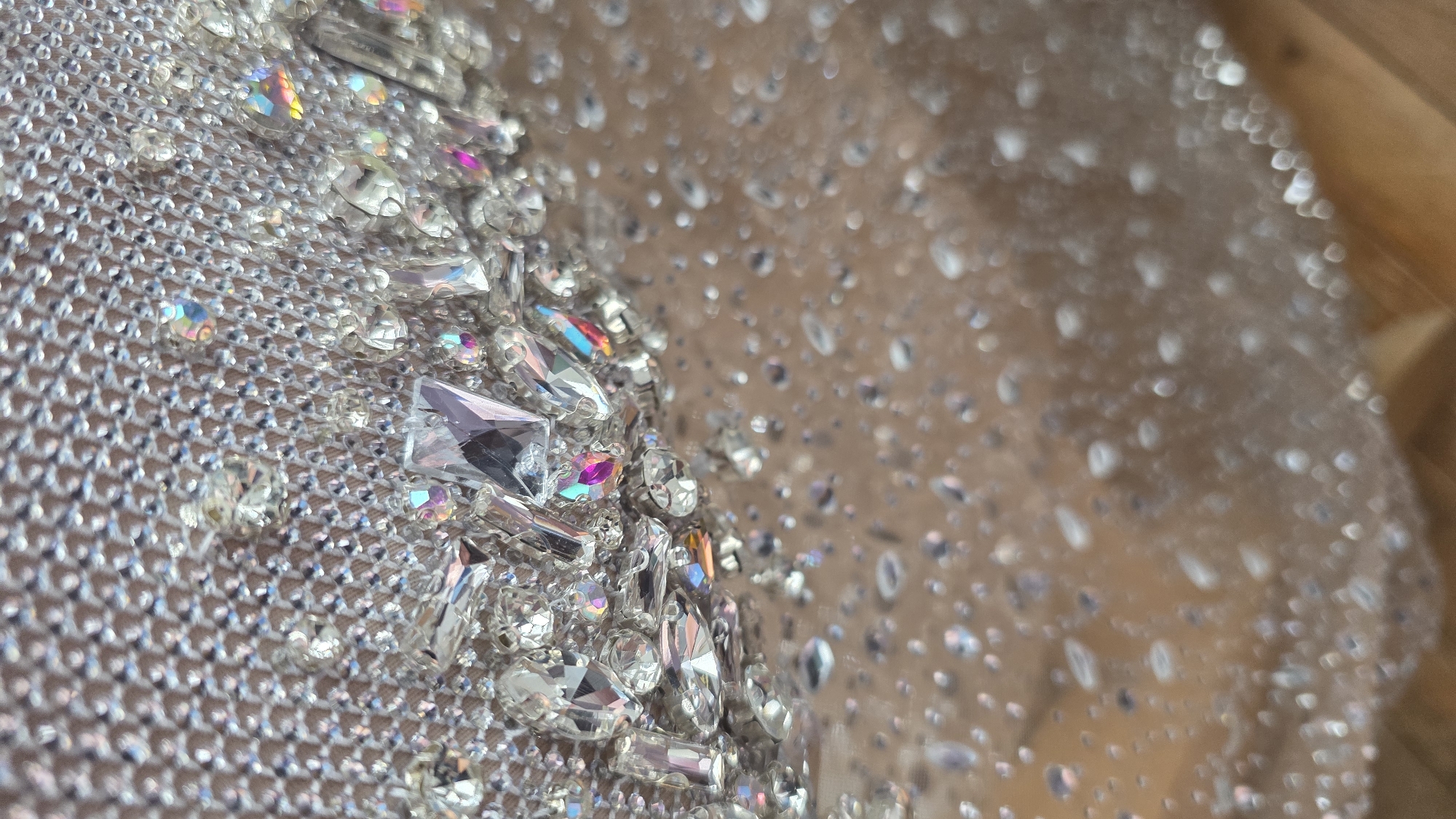 A close-up, high-angle shot captures a detailed view of an intricately embellished fabric or material. The left side of the frame, which is in sharp focus, showcases numerous sparkling gemstones, primarily clear and iridescent, with facets reflecting subtle hints of pink, blue, yellow, and green. These gems are of various shapes, including round, baguette, and marquise cuts, and appear to be securely attached to a finely woven, semi-transparent mesh fabric that itself exhibits a shimmering, metallic quality. A light brown or nude-toned surface is visible beneath the mesh.
As the eye moves towards the right side of the image, the focus softens, creating a bokeh effect where individual embellishments appear as diffused circles of light, suggesting a greater depth of field. The background on the right is a blurred, warm light brown or beige color, with faint hints of a lighter wooden surface towards the upper right corner. The lighting is bright, highlighting the reflective properties of the gems and fabric. No specific activity is taking place, as the image is a still life focused on texture and sparkle. There is no visible text or identifiable location cues within the image that would specifically indicate Blagoevgrad, Bulgaria. The overall impression is one of luxury, craftsmanship, and intricate detail.