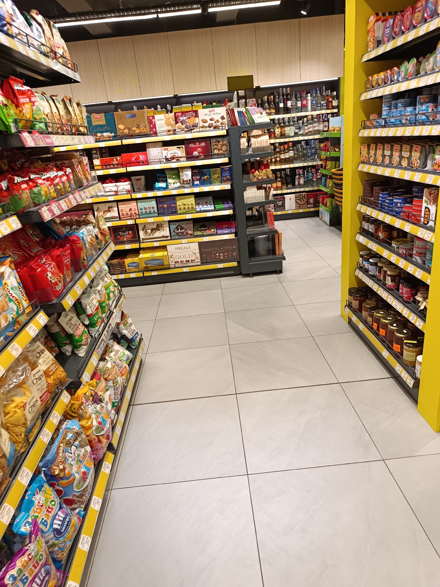 The image shows the interior of a grocery store, specifically an aisle stocked with various food products. On the left, shelves are filled with snack items like chips and crackers, while on the right, there's a yellow shelving unit stocked with items such as canned goods, condiments, and sauces. Further down the aisle, shelves display an assortment of chocolates, biscuits, and alcoholic beverages like liquor and wine. The floor is tiled in a light color, and the lighting appears to be fluorescent. There are no people visible in the image. The location is in the city of Unknown, Unknown.
