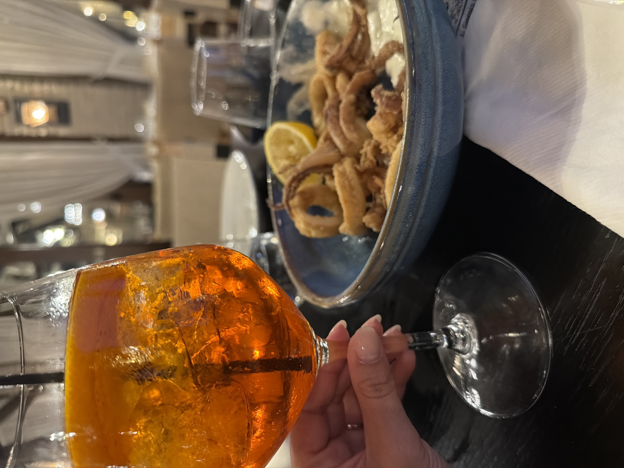 The image, rotated, captures a relaxed dining scene, likely in Makri, Greece. In the foreground, a person's hand, adorned with light-colored nail polish, holds a large glass filled with a vibrant orange beverage, consistent with an Aperol Spritz, containing ice cubes. The drink appears to have small bubbles, suggesting effervescence.
Positioned slightly behind and to the right of the drink is a blue ceramic bowl brimming with golden-brown fried calamari, accompanied by a fresh lemon wedge. A folded white napkin rests on a dark-colored table next to the bowl.
The background is softly blurred, showcasing a warm, ambient lighting scheme with multiple glowing light sources and elements of white, draped fabric. This setting suggests an indoor or covered outdoor area of a restaurant or bar, indicating the time of day is likely evening. The overall mood conveyed is one of enjoyment and leisure. There is no discernible text visible in the image.