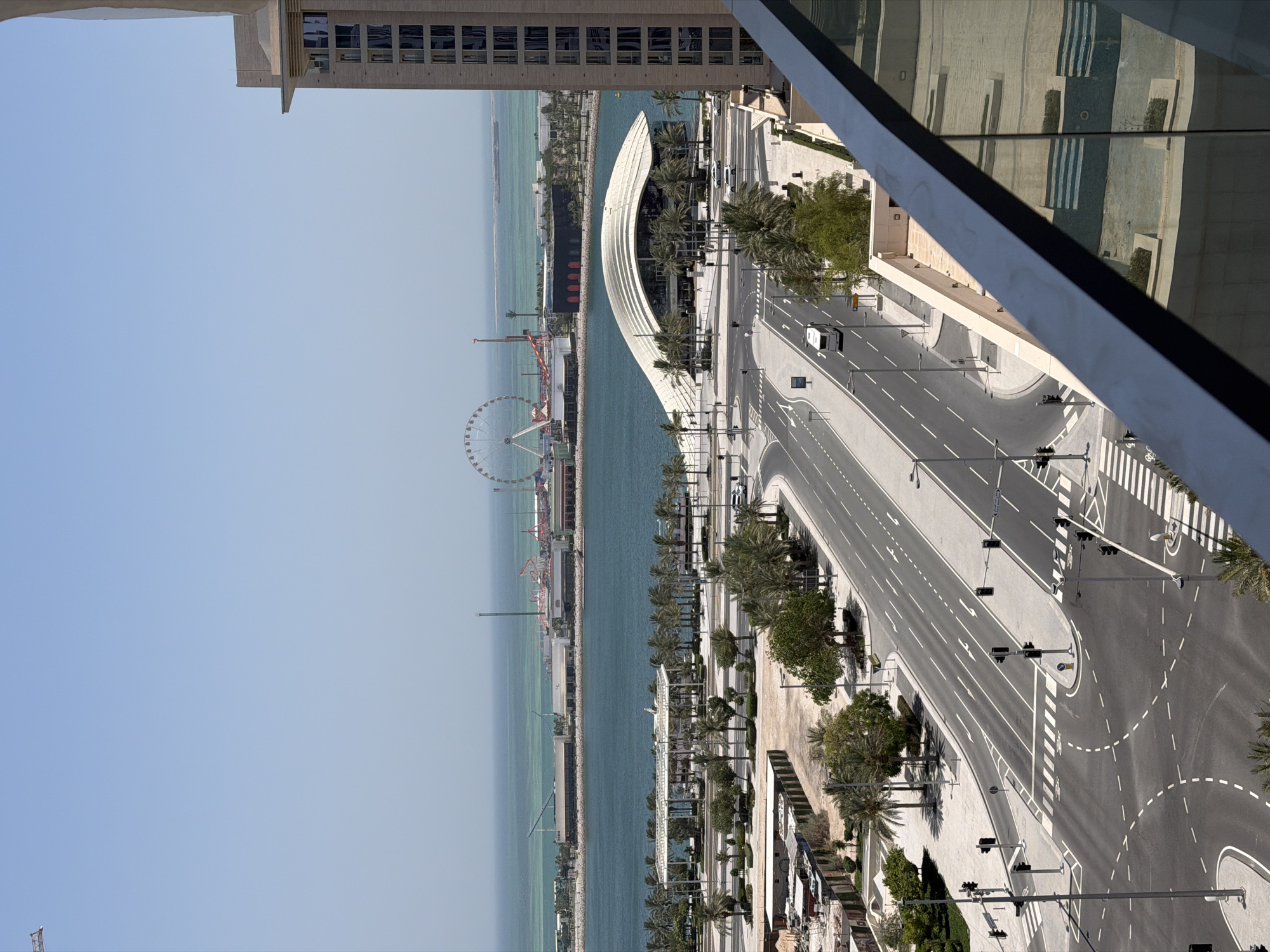 The image is a high-angle outdoor shot of a modern city scene with a body of water in the background. The foreground features a balcony railing with a reflection of a swimming pool. Below the balcony, a multi-lane road with clear white lane markings curves through the scene. Traffic lights are visible at intersections, and several vehicles, including a white bus and a few cars, are on the road. Pedestrian crossings are marked with white stripes.
On the left side of the road, there are construction sites with some scaffolding and building materials. Further back, there is a treed area with palm trees and other greenery. A large, distinctive white building with a curved roof is situated near the water.
Across the water, a waterfront area is visible, featuring what appears to be an amusement park with a Ferris wheel and other rides. Several buildings are also present along the waterfront. In the distance, the ocean stretches to the horizon under a clear blue sky. A tall, modern building with many windows is prominently positioned on the right side of the image. The overall impression is one of a well-developed, contemporary urban environment with recreational facilities by the water.