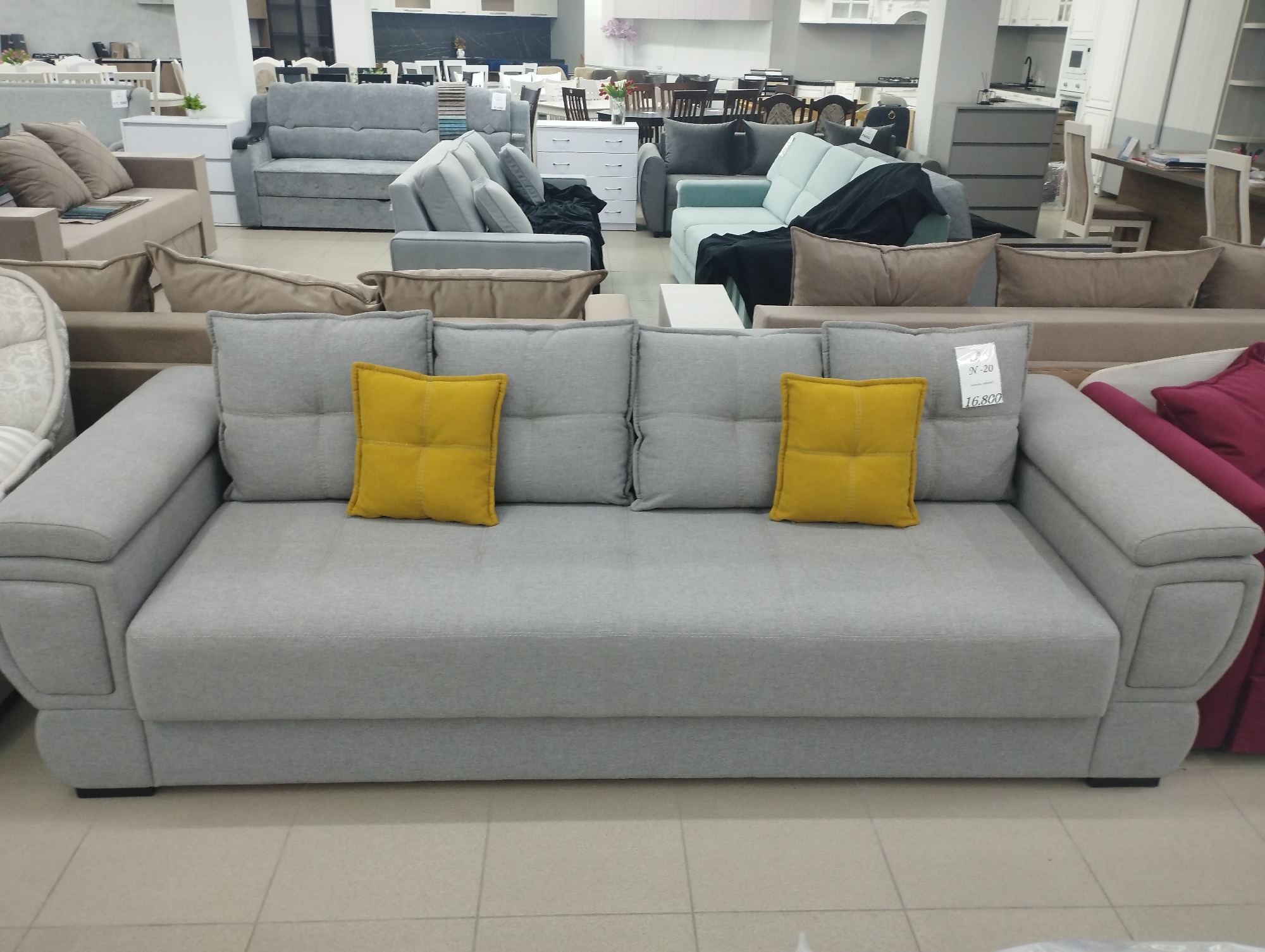 A light gray sectional sofa with two yellow throw pillows is prominently displayed in the foreground of a furniture store. The sofa is positioned on a tiled floor, and other furniture pieces, including more sofas and chairs, are visible in the background. The overall impression is that of a retail environment showcasing a variety of furniture. The store is located in Fălești, Moldova.