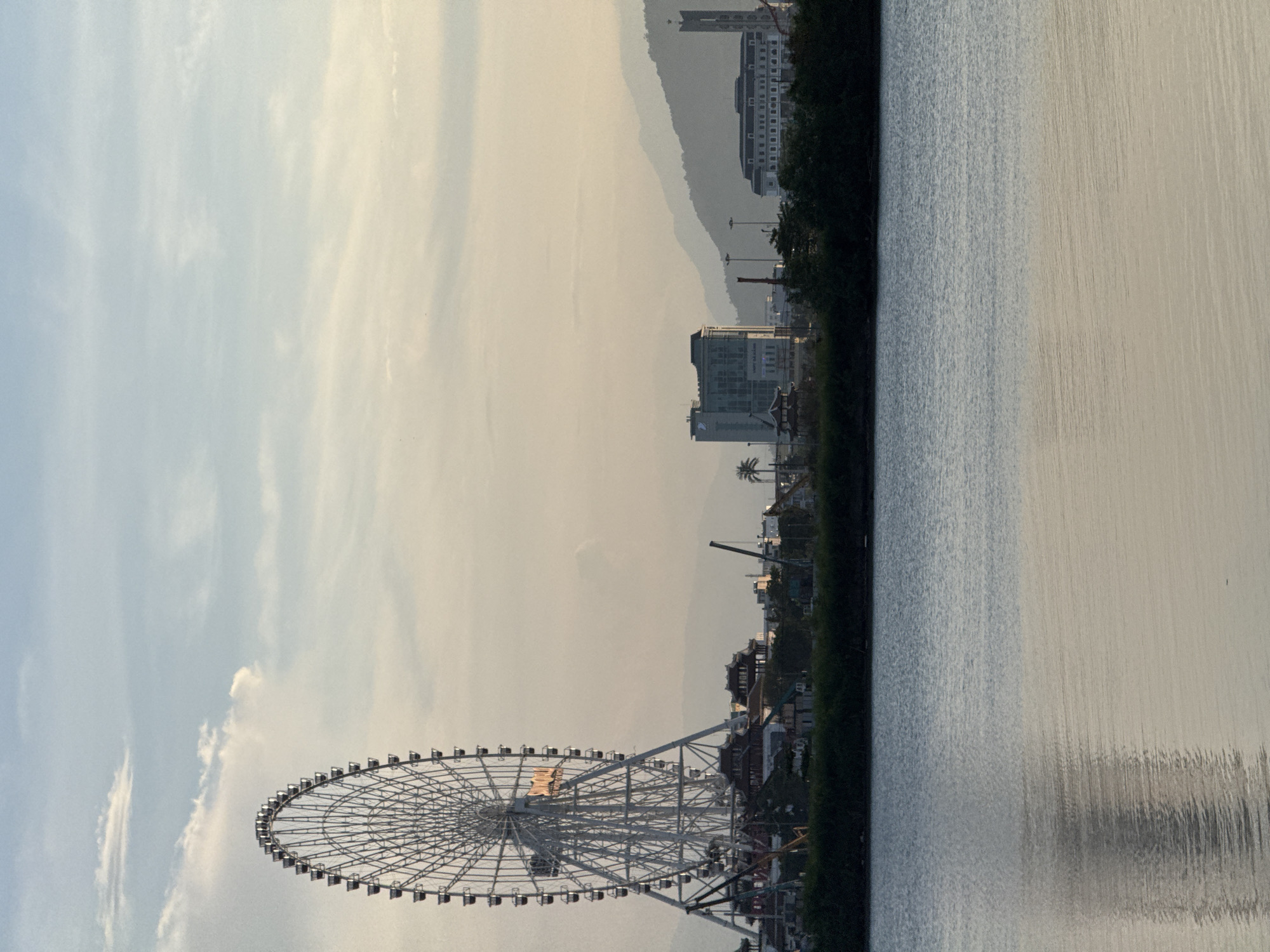 This image shows a wide shot of the city of Da Nang, Vietnam, at sunset. The foreground features a calm body of water, with ripples reflecting the sky. A dark green line of trees and bushes separates the water from the city's skyline. Dominating the left side of the frame is a massive Ferris wheel, its intricate metal structure silhouetted against the soft, hazy sky. The Ferris wheel is lit with orange lights, suggesting it is operational or recently illuminated. Below the Ferris wheel, various smaller structures, possibly part of an amusement park or entertainment complex, are visible. Further to the right, the city's architecture is apparent, with modern buildings and a more traditional-looking tower. In the background, layered, misty mountains recede into the distance, adding depth to the scene. The sky is a blend of pale blue and soft orange, typical of a sunset, with wispy clouds scattered across it. No people are visible in the image. The overall atmosphere is serene and picturesque, capturing the transition from day to evening in a coastal Vietnamese city.