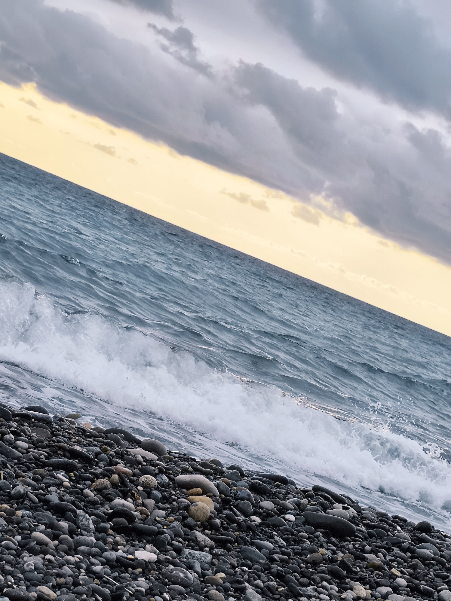The image captures a slightly tilted, close-up view of a pebble beach on the coast of Zemo Kvariati, Georgia. A wave is breaking onto the shore, with white foam and spray. The water is a deep blue-green, with ripples and texture. Above the water, the sky transitions from a pale yellow near the horizon to dark gray clouds. The beach is covered in a dense layer of smooth, dark-colored pebbles of varying sizes. There are no people or man-made structures visible in the frame. The overall impression is one of natural coastal scenery under an overcast sky.