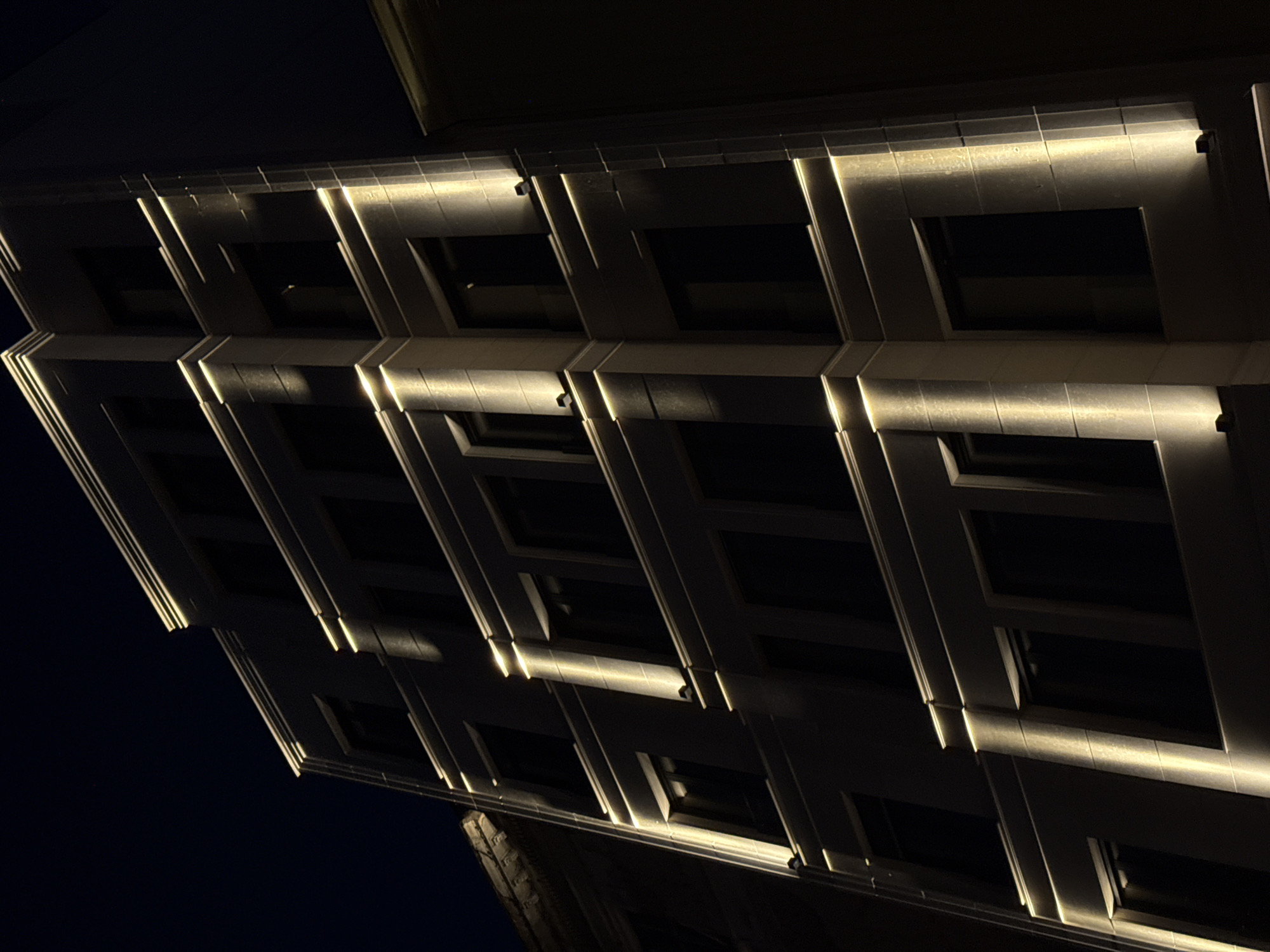 The image shows a portion of a building facade at night. The building is illuminated with linear lights, emphasizing the architectural details such as the window frames and cornices. The windows are dark. The angle of the shot looks upwards toward the building. The city is Belgrade, Serbia.