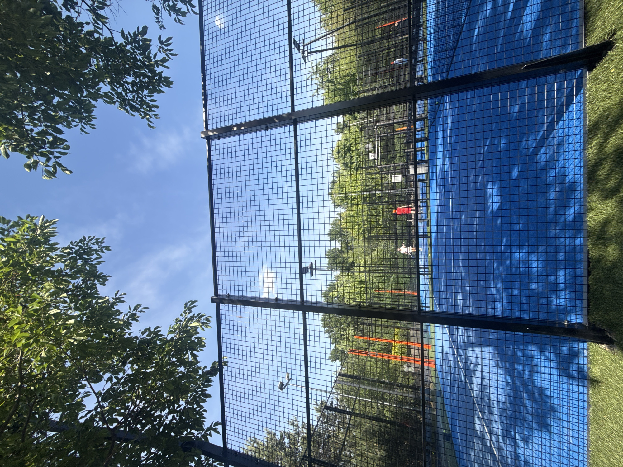 The image captures an outdoor padel court in Nemchinovo, Russia, viewed from a low angle through a black wire mesh fence. The fence, composed of square grids and prominent vertical support beams, occupies the majority of the foreground and casts distinct, intricate shadows across the blue playing surface. White lines demarcate zones on the court.
Two individuals are visible on the court. On the left side, a person wearing a red shirt is partially obscured by a black court structure and the fence. On the right, further back on the court, another person in a light-colored (possibly white or beige) top and shorts is visible, appearing to hold a racket, with a yellow ball present on the court near their position.
The court is enclosed by additional sections of black mesh fencing and surrounded by dense, tall green trees, which form a natural backdrop. Several tall black poles equipped with floodlights are positioned around the court for nighttime illumination. A striking orange, upright structure is visible in the background through the trees on the left.
Above the court and trees, a bright, clear blue sky with a few scattered, wispy white clouds is visible. Lush green tree foliage frames the top edges of the image. The immediate foreground, beneath the blue court, consists of a strip of bright green artificial grass. The scene is illuminated by strong sunlight, indicative of a clear day.