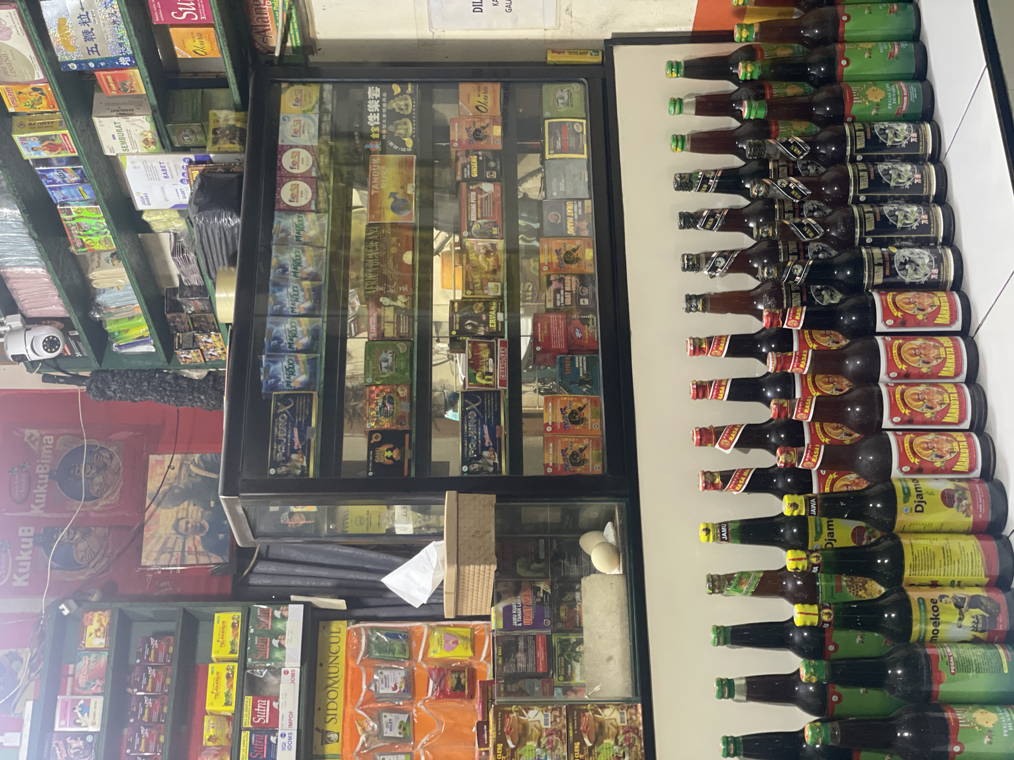 The image shows the interior of a shop in North Jakarta, Indonesia. A glass display case is stocked with various health products and supplements. Bottles are lined up on a white surface next to the case. Some bottles bear the labels "Djam," "Moekoe," and "Pegal Linu Husada," while boxes display brands such as "Sutra," "Sidomuncul," and "Okura." The shelves behind the display case are also filled with similar items. Posters and advertisements are visible on the walls. A security camera is mounted on the wall. The items are densely arranged.