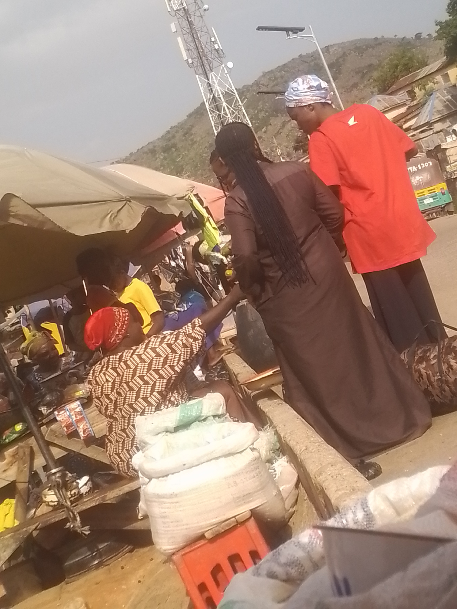 The image depicts a market scene with several people. In the foreground, a person in a patterned brown and white garment and a red headscarf is partially visible, appearing to be engaged in an activity. Behind them, a person with long dark braids, wearing a dark brown dress, stands looking away. To the right, a person in a bright red t-shirt and dark pants is also visible.
The setting appears to be an outdoor market, indicated by the presence of stalls with awnings and various goods displayed, including large sacks of what look like grains or produce, and what could be packaged items. In the background, there is a telecommunications tower and a hillside with some buildings. A vehicle, possibly an auto-rickshaw, is also partially visible on the right side of the frame.
The lighting suggests it is daytime, likely sunny given the shadows. The overall atmosphere seems active and busy, typical of a marketplace. There is a piece of text visible on the side of the vehicle that reads "TA 1307". The image captures a moment of commerce and social interaction in a bustling environment.