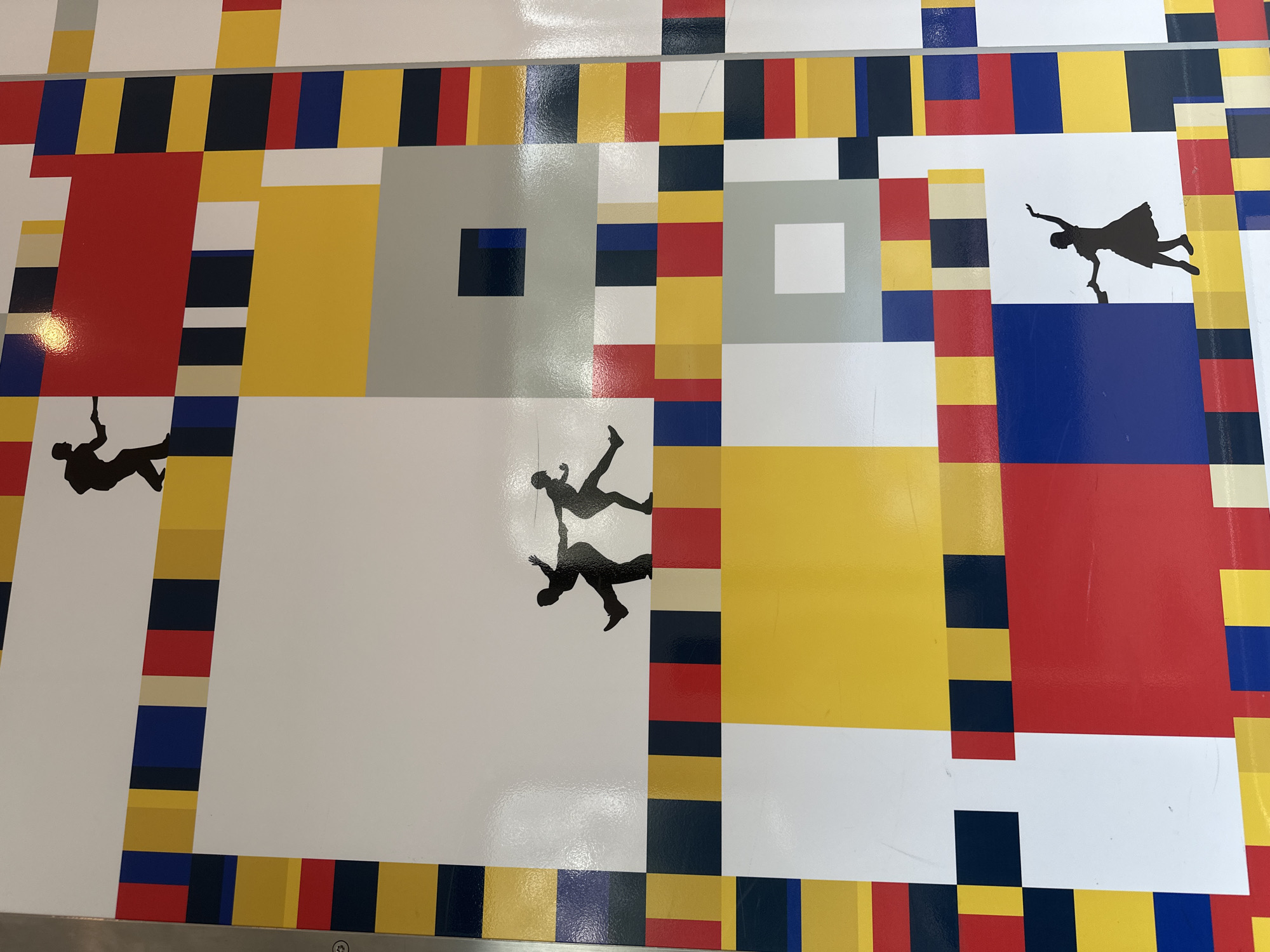A high-angle shot depicts a section of a large, flat surface adorned with a design reminiscent of Piet Mondrian's abstract style, featuring rectangular blocks of red, yellow, blue, white, gray, and black. The design is not a perfect replication of Mondrian's work, as it includes narrower stripes of color framing larger rectangular areas.
Superimposed onto this geometric background are several black silhouettes of people in acrobatic poses or in the act of falling. One silhouette appears to be dangling by an arm. The image's location is in Best, Netherlands.