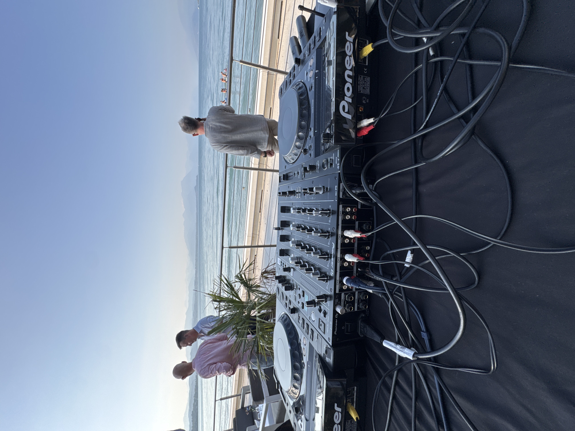 The image depicts an outdoor scene at a waterfront location in Desenzano del Garda, Italy. In the foreground, a Pioneer DJ setup is prominent, featuring two turntables and a mixer, with numerous cables connected. In the midground, two men in casual attire stand near a railing, facing away from the camera, with a body of water and distant mountains in the background. A man in a grey shirt is also visible, facing the water and holding a phone to his ear. Further in the background, several individuals are seen swimming in the water. The environment suggests a relaxed daytime atmosphere.