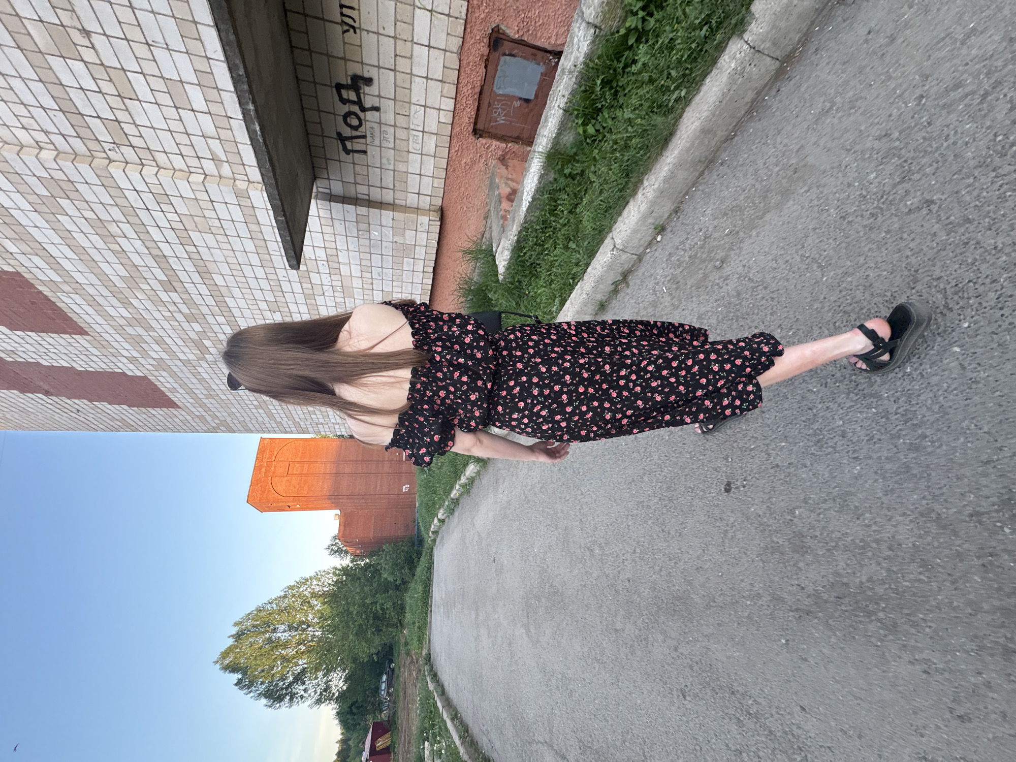 A girl with long brown hair, wearing a floral print dress and black sandals, is walking on a gray asphalt street. To her left are trees and a brick building in the distance, under a clear blue sky. To her right are concrete curbs lined with grass and a building with white tiled walls and an orange wall with a boarded-up window. There is graffiti on the white wall. This is a street scene in Izhevsk, Russia.