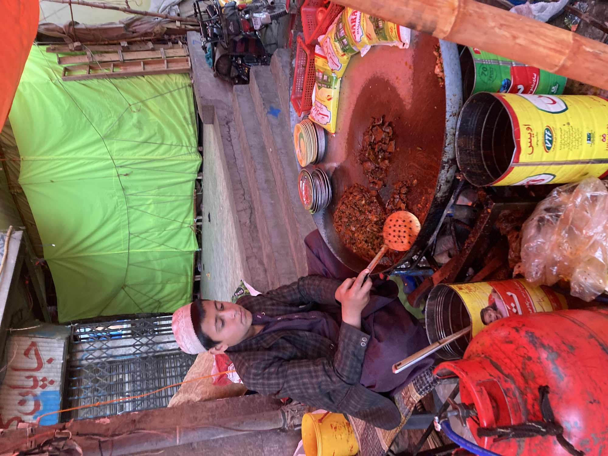 An overhead-angle image depicts a young male preparing food at a street stall in Quetta City Tehsil, Pakistan. The individual, dressed in a plaid jacket, a dark purple salwar kameez, and a white skullcap, sits cross-legged on a mat, actively stirring a mixture of cooked meat and vegetables on a large, circular, flat griddle with a slotted metal spatula. To his left, a red LPG cylinder with a blue hose powers the cooking apparatus. Two large, repurposed yellow metal cans stand near the griddle, one labeled "اويس VTF" with a girl's image. Stacks of ceramic and metallic plates, red plastic baskets, and several yellow plastic bags (one featuring "ZARA BASMATI RICE" branding) are arranged near the griddle for serving. In the background, concrete steps lead up to an area partially enclosed by a large green tarpaulin. A dark motorcycle is parked beside the steps. Further left in the background, a shop front with a pulled-down metal security shutter displays "جاپان" (Japan) in red Urdu/Arabic script. An orange fabric canopy partially covers the scene from above. A yellow plastic bucket is on the far left.