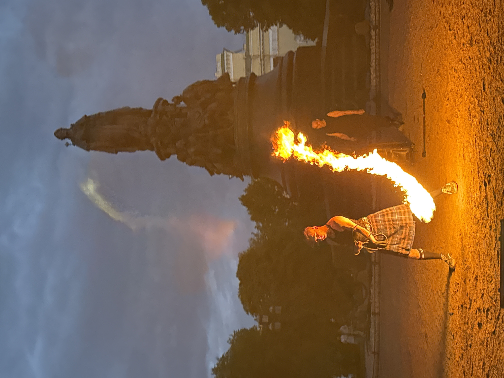 The image depicts a nighttime scene on Palace Square in Saint Petersburg, Russia, in front of the Alexander Column. A performer in a kilt is engaged in fire-spinning, with a large jet of flame erupting from a device he is holding. The flame is bright orange and yellow, casting a warm glow on the ground and the performer. Behind the performer, another individual in a dark outfit stands watching. The Alexander Column, a prominent monument, is visible in the background, silhouetted against the dusky sky. Trees and parts of buildings can also be seen on the periphery of the square. The sky is overcast with dark clouds.