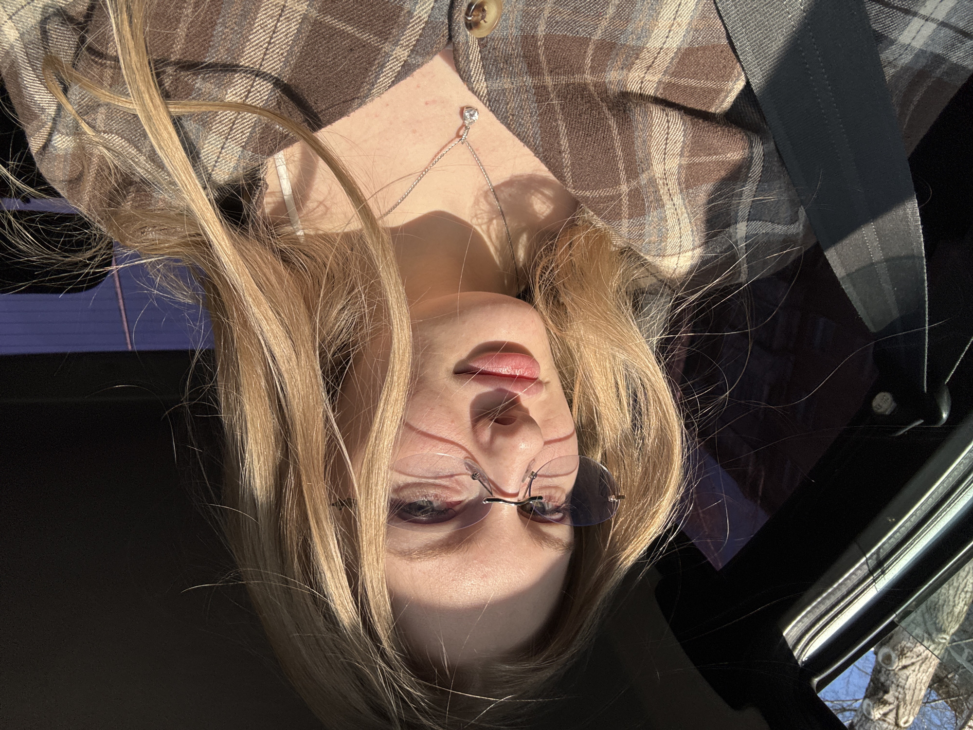 A selfie of a light-skinned woman with blonde hair wearing rimless tinted glasses and a silver necklace with a pendant. She is wearing a brown and gray plaid button-down shirt with a single button visible. A dark gray seatbelt is visible diagonally across her upper body. The photo appears to have been taken inside a car, with the interior roof and window visible. A tree and blue sky are seen through the window. I am unable to place the image in the city of Perm, Russia.