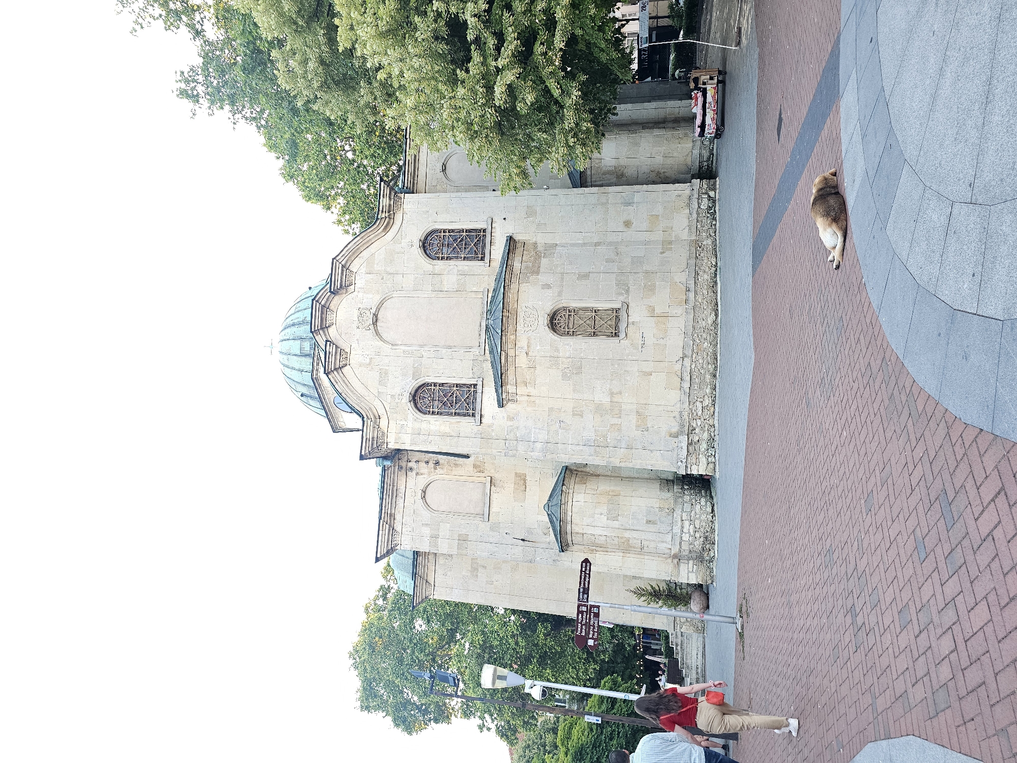 An exterior shot captures a pale stone building with a turquoise dome in the city of Unknown, Unknown. The building, appearing to be a church or mosque, features arched windows with intricate metalwork. Trees with lush green foliage surround the structure. In the foreground, a paved walkway with a red brick pattern is visible. A couple walks away from the camera on the left, and a dog is lying down on the pavement in the lower right portion of the image. A street signpost with directional arrows stands near the couple. A mobile food cart is parked to the right of the building. The sky is overcast and bright.