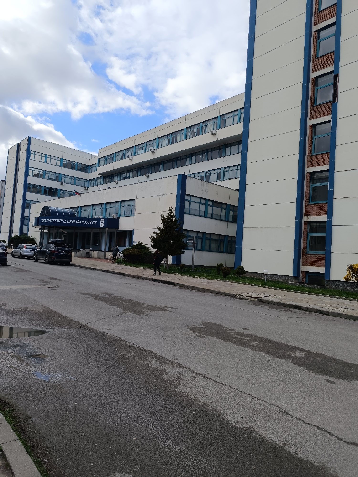 The image shows the Elektrotehnički Fakultet (Faculty of Electrical Engineering) building in Sofia, Bulgaria. The building is a large, modern structure with white and blue accents. There are multiple cars parked in front of the building, and a few people are walking on the sidewalk. The street in front of the building is wet, with puddles visible. The sky is cloudy, suggesting a recent or ongoing rain.