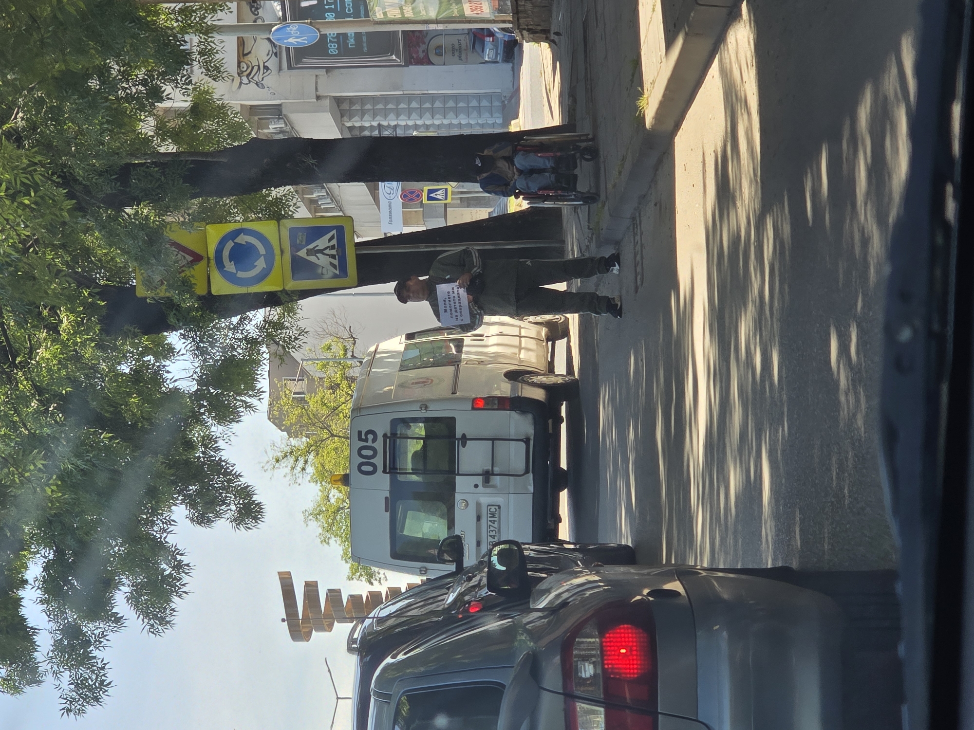 The image shows a street scene, likely in a city, with a white van marked "005" and the license plate "B 4374 MC" parked on the side of the road. A man stands next to the van holding a sign written in a Cyrillic script that says "Моля, помогнете на детето с операцията" which translates to "Please help the child with the operation". The man is wearing a tracksuit. Further down the sidewalk, a person is sitting in a wheelchair. Traffic signs indicating a roundabout and pedestrian crossing are visible. A gray car with its brake lights on is stopped behind the van. Buildings and trees line the street. Part of a decorative sculpture is visible.