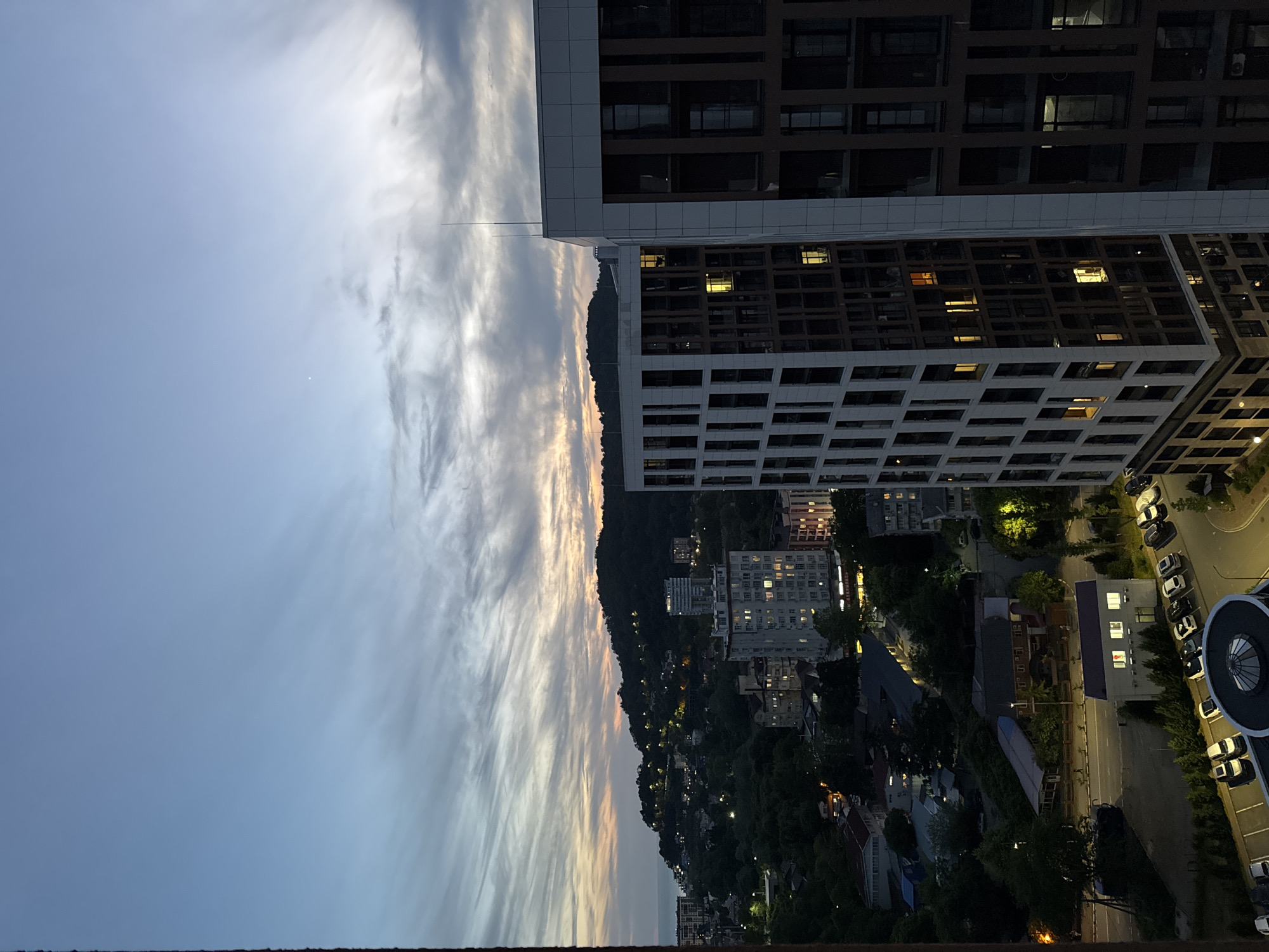 The image is a high-angle, medium shot of a city landscape in Vardane Microdistrict, Russia, during dusk. The sky is a gradient of pale blue and muted orange, with wispy clouds stretched across it, indicating the end of the day. In the foreground, two modern high-rise buildings dominate the right side of the frame. The building on the left has a white facade with a grid of dark windows, some of which are illuminated with a warm yellow light. The building to its right has a darker facade, with a similar grid of windows, also showing scattered interior lights.
Beyond the immediate buildings, the city sprawls out towards a dark, tree-covered hill in the background. Numerous smaller buildings and houses are scattered across the hillside, many with illuminated windows, suggesting residential areas. Streetlights dot the landscape, casting pools of light on roads and pathways. The sea is visible on the far left, meeting the coastline where more buildings are clustered. Trees and vegetation are abundant, creating a sense of a city integrated with nature. The overall impression is of a developing urban area at twilight, with the lights of the city starting to glow against the darkening sky and natural landscape. There are no people visible in the image.