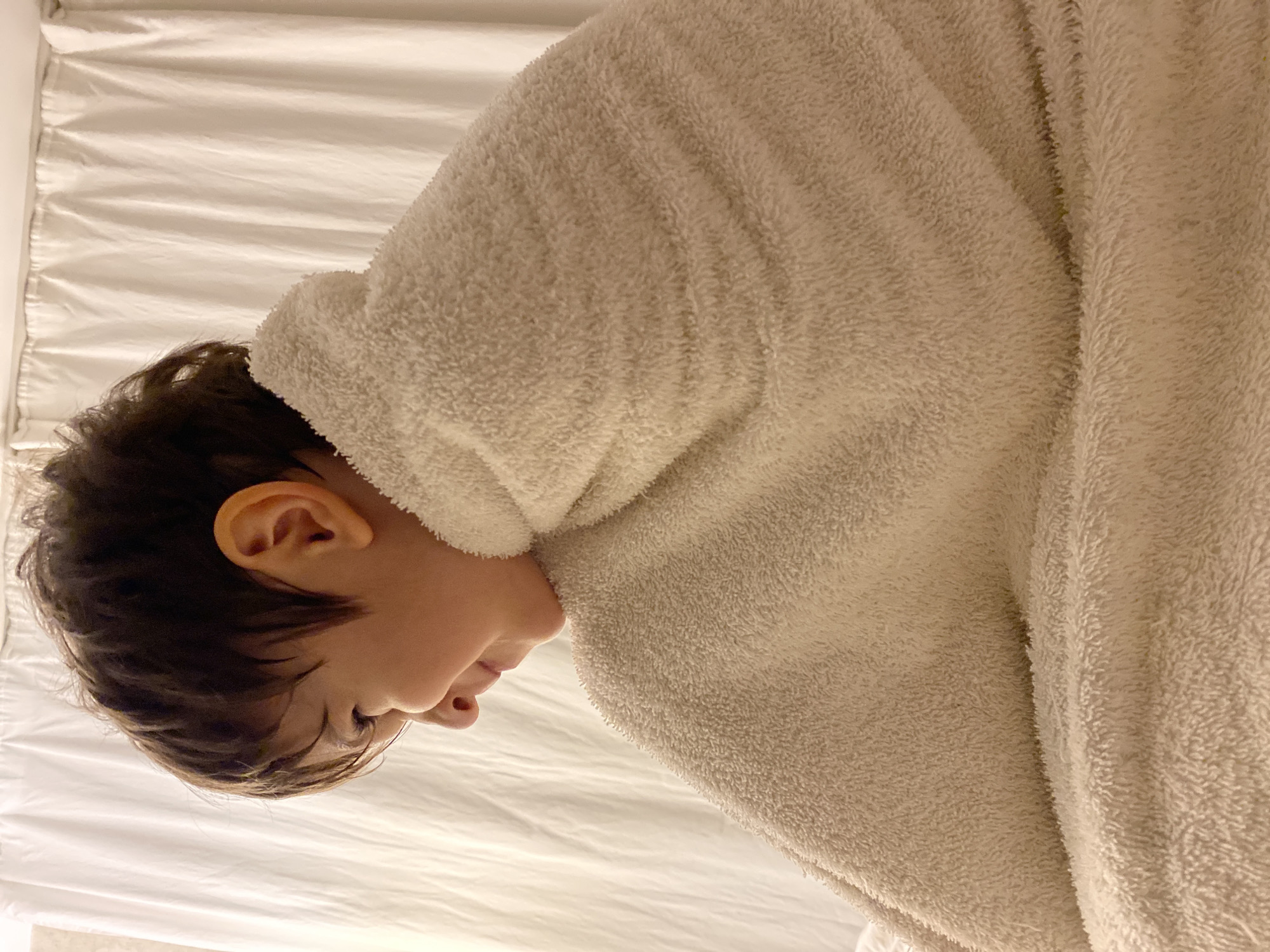 A close-up shot of a child lying on a white bed, partially covered by a white, plush robe. The child's head is visible, with dark hair, an ear, and a profile view of their face. The robe covers the majority of their body. The photograph is taken from a high angle. There is no indication of the location being Yekaterinburg, Russia, or any specific city.