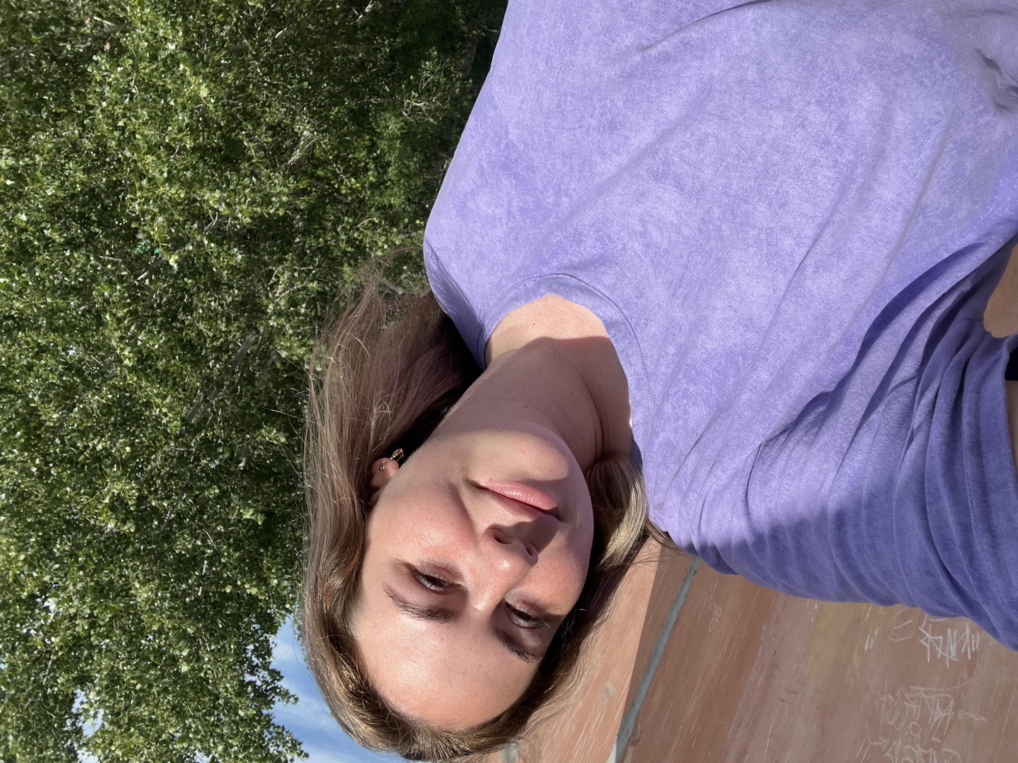The media file is a portrait of a young woman in an outdoor setting. She has blonde hair and is wearing a purple t-shirt. Her face is turned slightly towards the camera, and she has a gentle smile. Her eyes are brown and appear to be looking slightly downwards. She is wearing a gold earring with a heart-shaped charm. The background shows lush green trees and a bright blue sky, suggesting it is daytime with clear weather. There is a brick-like structure in the foreground, which appears to be part of a wall or ramp, with some graffiti visible on it. The overall impression is one of a relaxed and pleasant outdoor moment.