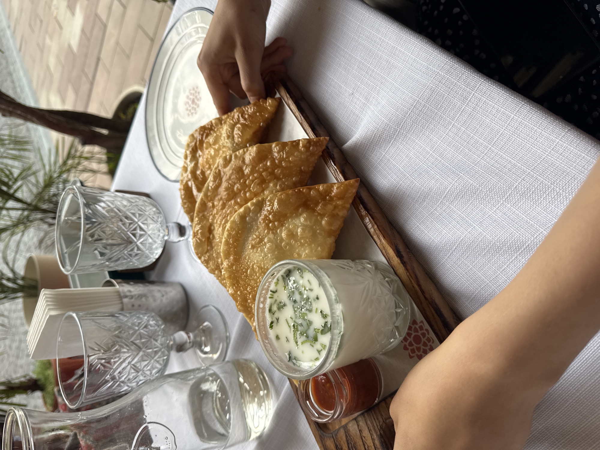 A hand reaches to pick up a golden-brown, crescent-shaped pastry from a wooden tray on a white tablecloth. The pastry appears to be fried and flaky, with a slightly irregular, puffed surface. Three similar pastries are already on the tray. Next to them, a small glass jar contains a white, creamy liquid with visible green herbs stirred in, possibly a yogurt-based drink. Below this, a tiny glass jar holds a reddish sauce, likely a condiment. The scene is set at a table, suggested by the tablecloth and glassware, which includes several ornate, cut-glass goblets and a clear glass bottle. In the background, a white plate with a subtle pink pattern is visible, along with glimpses of greenery and a building's facade, hinting at an outdoor or well-lit indoor setting. The overall impression is that of a meal or snack being served, possibly in a restaurant or a home dining area. The lighting suggests daytime. The image is oriented vertically, as if taken from a slightly elevated angle.