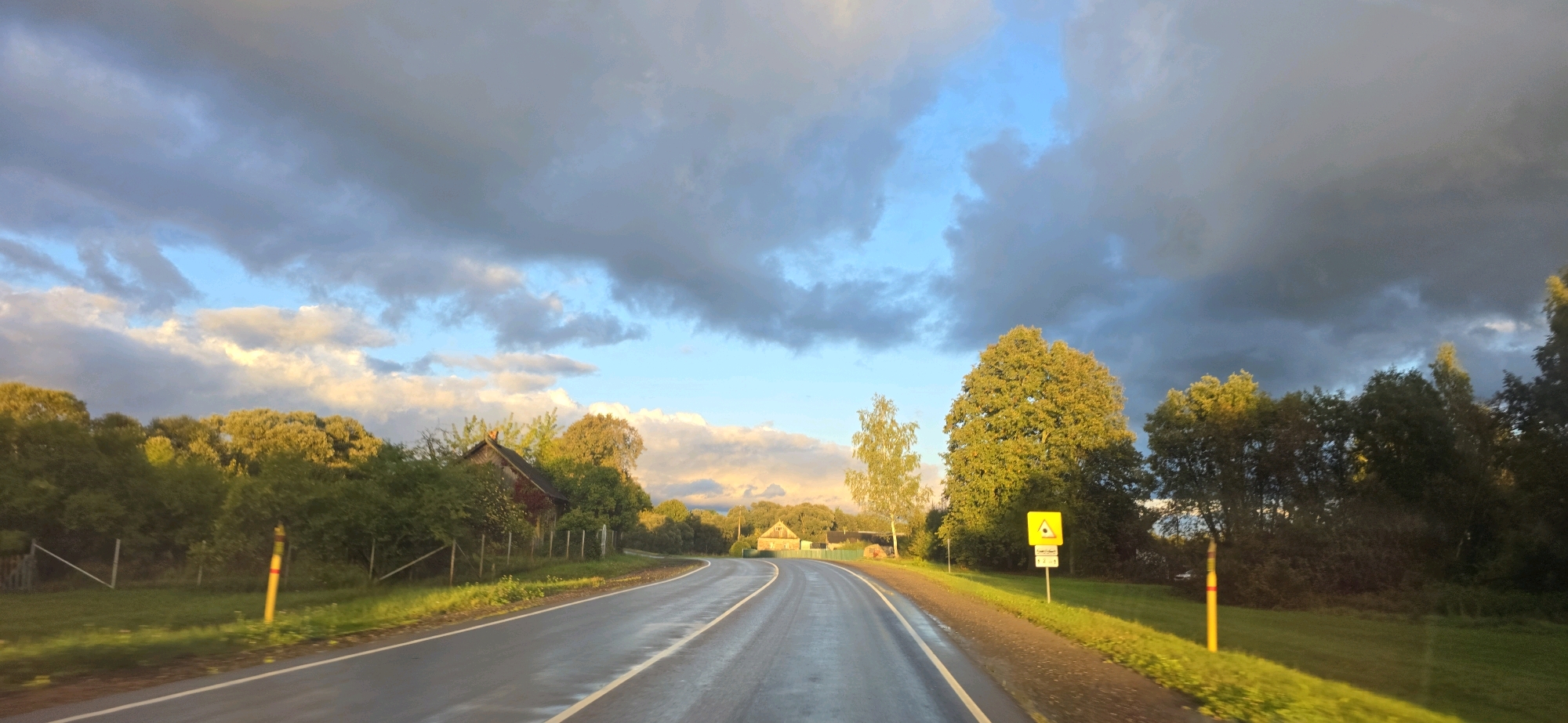 The image captures a scenic drive through what appears to be a rural area in the vicinity of Фарынаўскі сельскі Савет, Belarus. A paved, two-lane road, wet and reflective, curves gently to the right, bordered by lush green grass and trees. The roadside is marked by yellow posts with red and yellow reflective bands. On the left side of the road, a rustic wooden building with a dark roof is nestled among trees, and a fence runs along the property line. Further down the road on the right, a yellow triangular warning sign with a black symbol is visible. The landscape is characterized by dense foliage and scattered trees, with a few houses visible in the distance. The sky is dramatic, filled with a mixture of dark, brooding clouds and patches of bright blue sky, with sunlight breaking through in places, casting a warm glow on the trees. The overall impression is of a late afternoon or early evening scene, possibly after a rain shower.