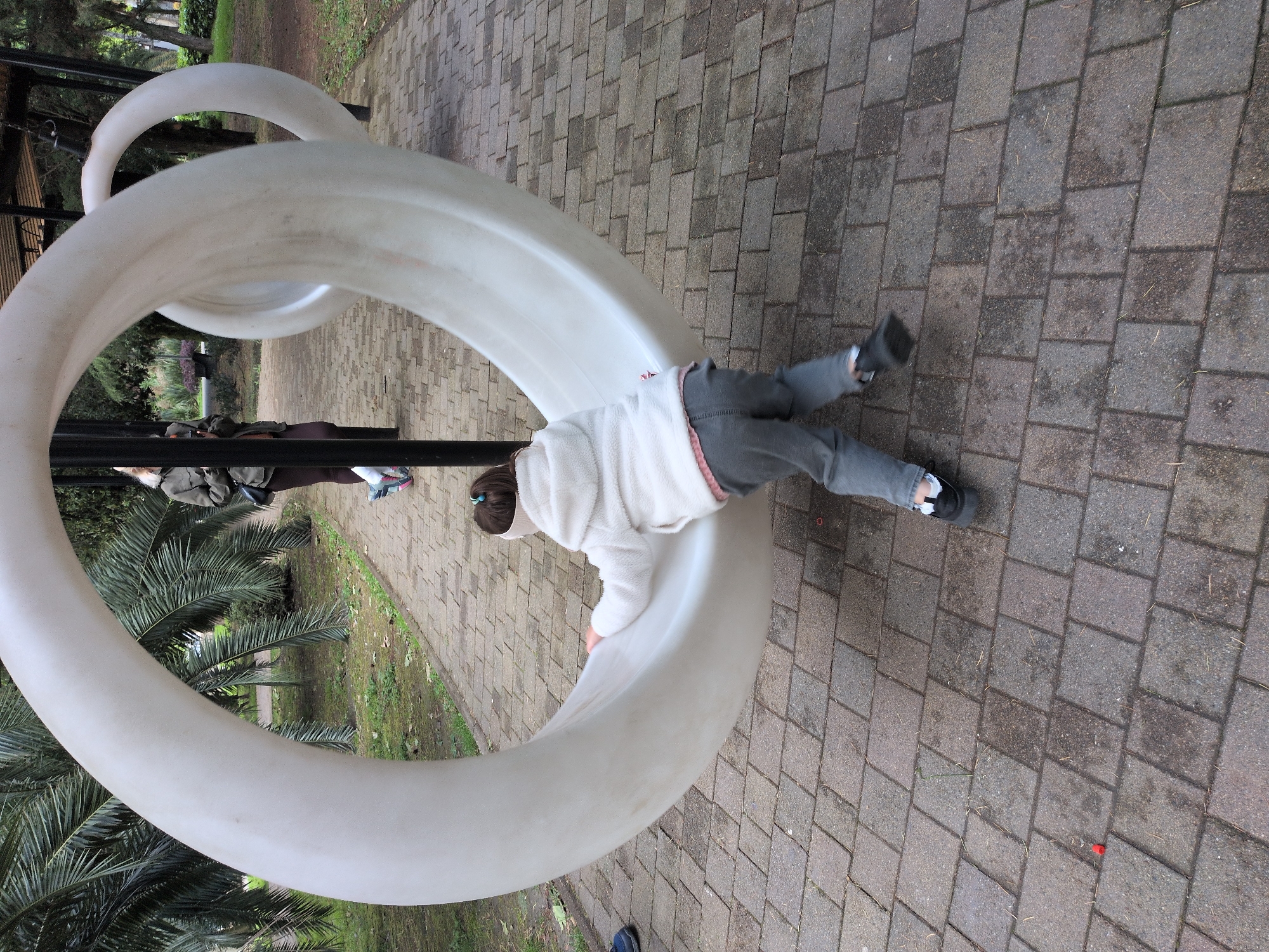 A child is crawling through a white, ring-shaped playground structure on a brick-paved surface. The child is wearing a white jacket and gray pants with black shoes. In the background, there is a green space with palm trees and a covered structure. The location may be in Sochi, Russia.