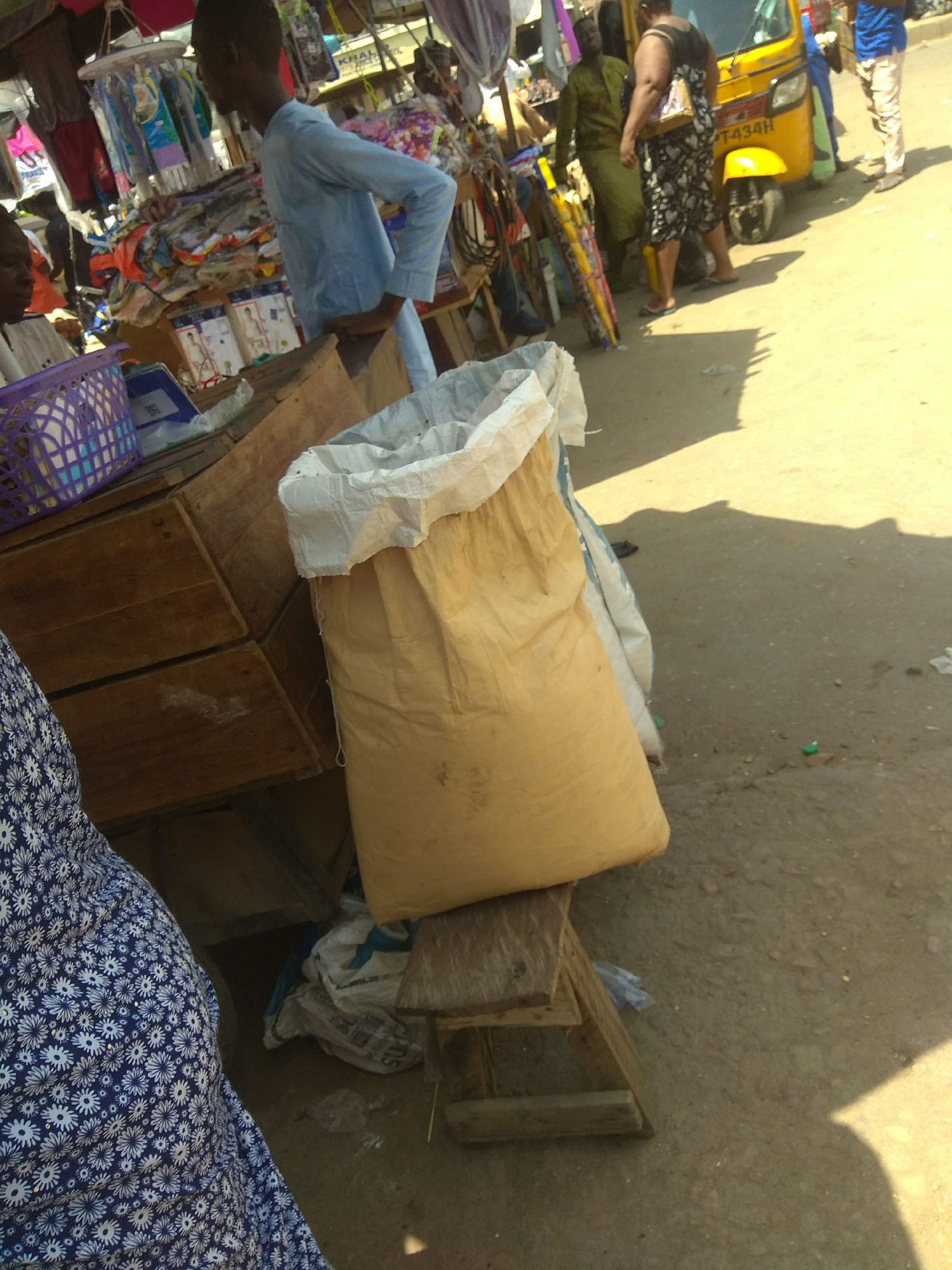 In Jalingo, Nigeria, an outdoor market scene unfolds with a narrow focus on a stall and its immediate surroundings. A young Black male, wearing a light blue long-sleeved shirt and dark trousers, stands behind a wooden stall laden with various goods, including what appears to be clothing items hung on display. To the left, a purple woven basket is visible on the stall. In front of the stall, a large, light brown burlap sack with a white opening is prominently positioned, resting on a wooden structure. A portion of a person's blue and white floral patterned garment is visible in the lower left corner, suggesting another individual is present. In the background, a yellow motorized tricycle, commonly known as an "okada" or "keke napep," is parked. Several other individuals are also visible, some standing near the tricycle and others further in the background, engaged in market activities. The ground is paved with dirt or gravel, and the scene is bathed in bright sunlight, casting distinct shadows. The overall impression is one of a bustling, everyday market in a Nigerian city.