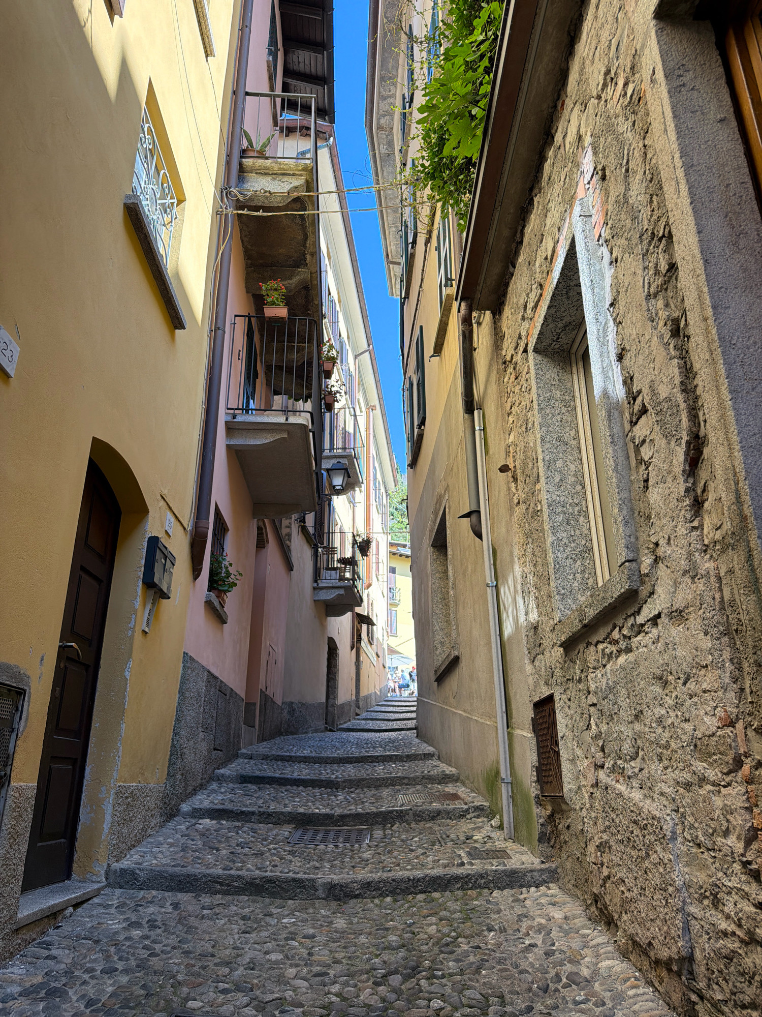 The image depicts a narrow, cobblestone alleyway in San Giovanni, Italy, leading uphill between two buildings. The building on the left is painted a pale yellow and features a dark wooden door with an arched top, a mailbox, and a window with decorative white grilles. The building on the right is constructed of rough, textured stone and has a large, gray-speckled window frame. Several balconies with wrought-iron railings are visible on the left building, some adorned with potted plants. The alley is lined with steps made of large, rounded cobblestones. In the distance, at the top of the alley, a few people can be seen walking, and the sky is a clear blue. The scene is bathed in sunlight, casting shadows on the buildings and the alleyway.