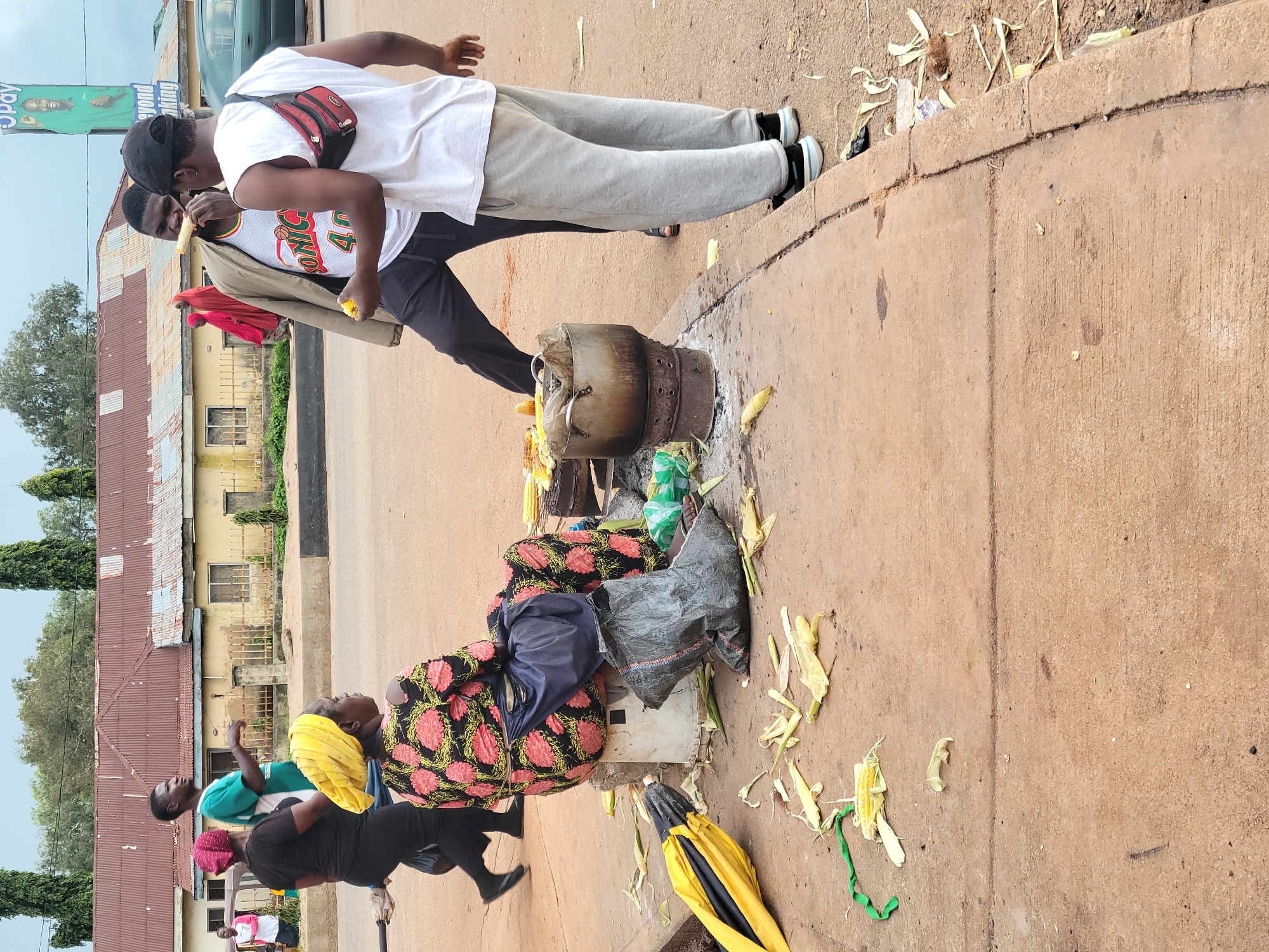 In the city of Gura Top, Nigeria, a street vendor is seen selling roasted corn. A woman wearing a black dress and a pink headscarf is seated on a white stool, her back to the camera, wearing a colorful pink and black floral dress and a yellow headscarf. A large black bag is behind her, and an open yellow umbrella is on the ground to her left. In front of her, a pot is heated over a makeshift stove, filled with corn. Two young men are standing, one of whom is eating a piece of corn. The ground is littered with corn husks and debris. In the background, buildings and a billboard advertising "Pay Beyond Banking" are visible, along with a few trees. The scene depicts a typical street market setting.